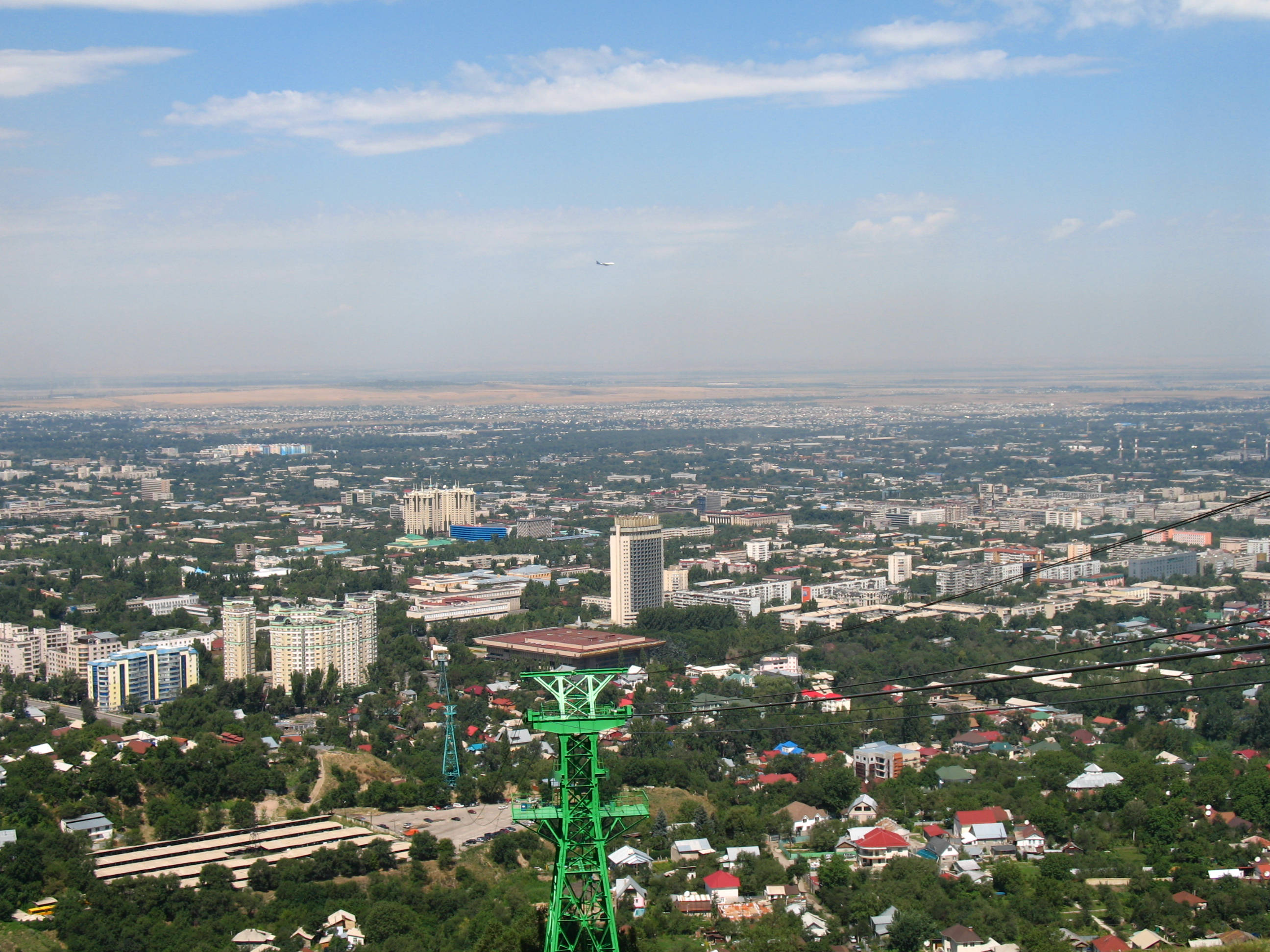 Kazakhstan 桌布 壁紙 — 廣闊城市景觀的全景視圖，在明亮的藍天下有散落的雲朵，前景有一個突出的綠色纜車塔。