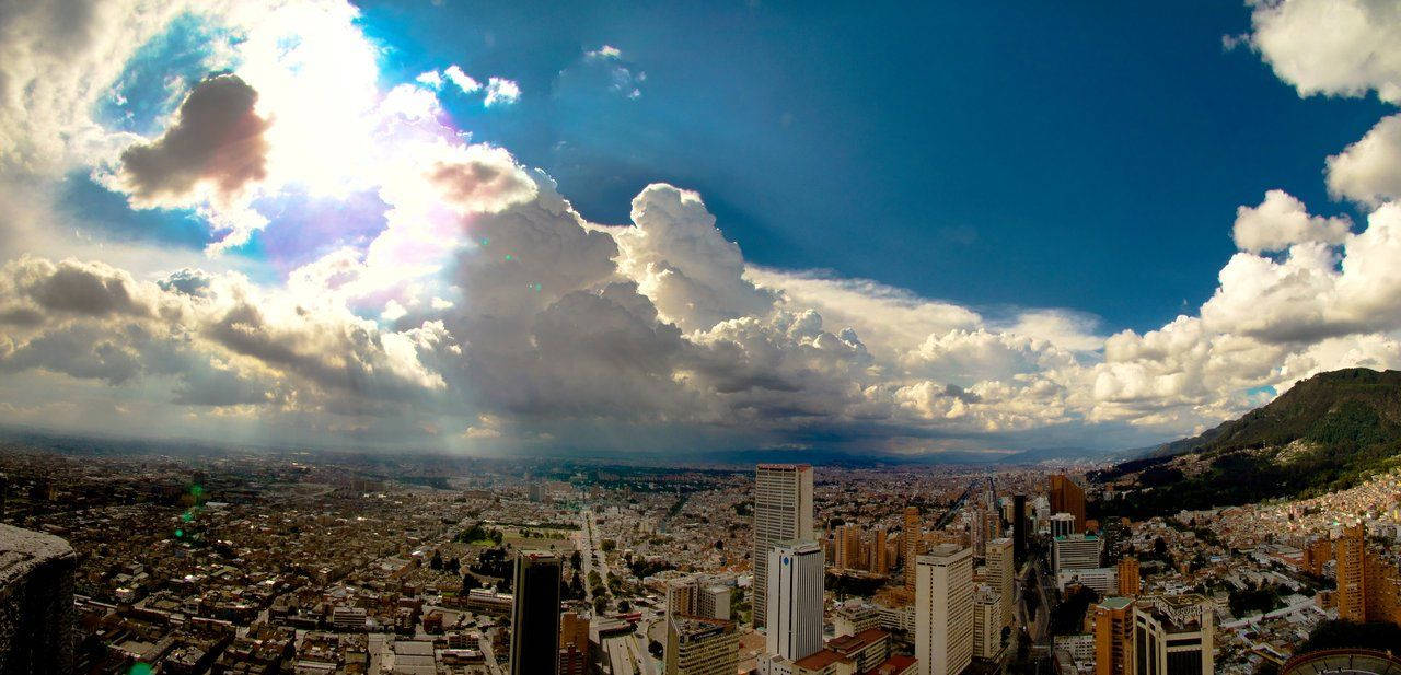 Bogota 桌布 壁紙 — 廣闊城市景觀的全景視圖，戲劇性的天空下，陽光穿過蓬鬆的白雲。