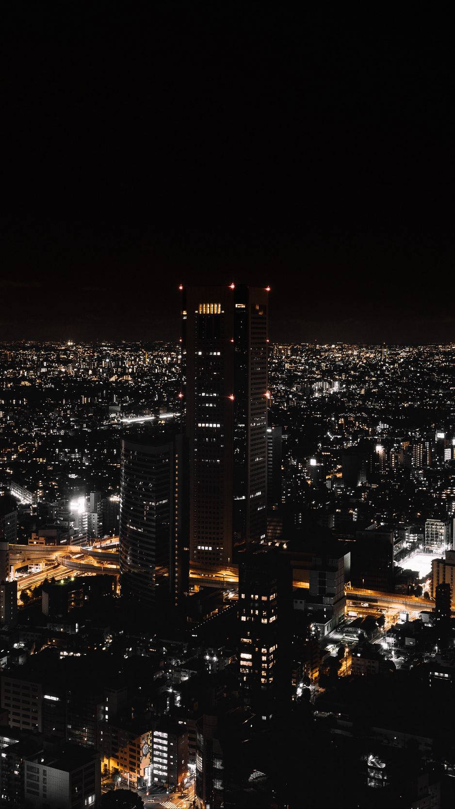 桌布 壁紙 — 廣闊城市景觀的夜間鳥瞰圖，以一座高聳的摩天大樓為主，在漆黑的天空中散發著溫暖的光芒。