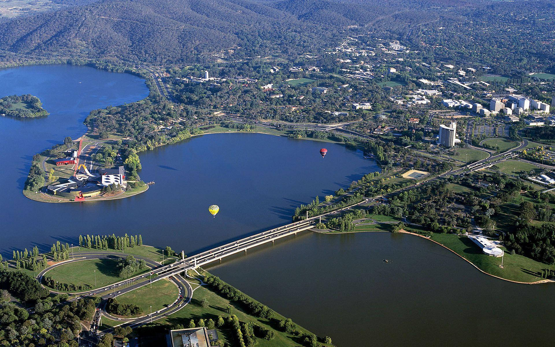 Canberra 桌布 壁紙 — 廣闊城市景觀的鳥瞰圖，蜿蜒的道路、橋樑和湖泊被綠色山丘和漂浮的熱氣球環繞。