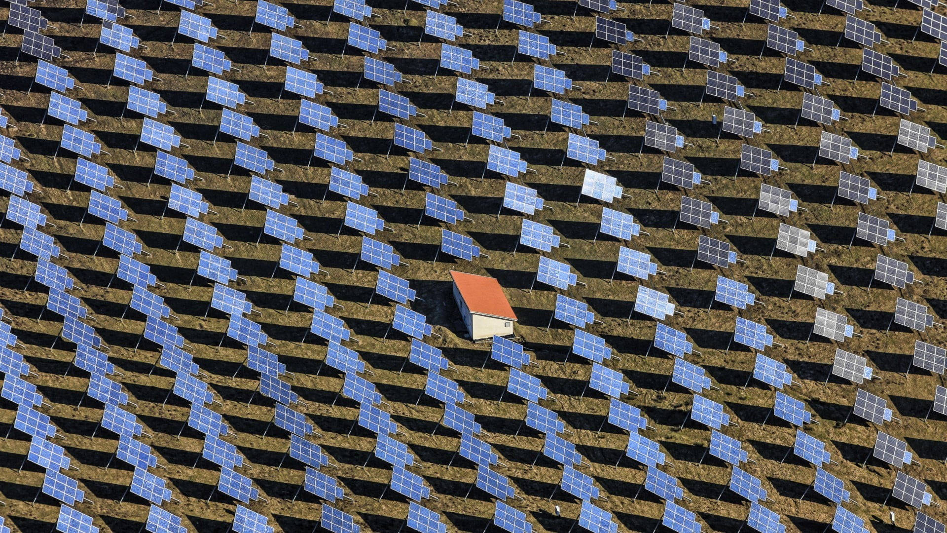 Home 桌布 壁紙 — 廣闊太陽能發電場的鳥瞰圖，藍色太陽能板以對角線排列，圍繞著一座紅色屋頂的小建築。