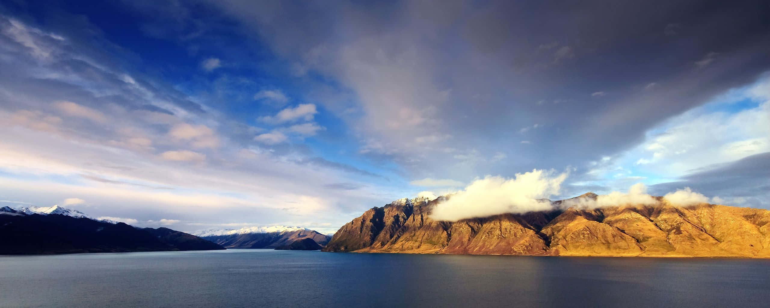 雙螢幕 桌布 壁紙 — 廣闊寧靜的湖泊映照著戲劇性的天空，金色光芒灑落在部分被雲霧遮掩的崎嶇山脈上。