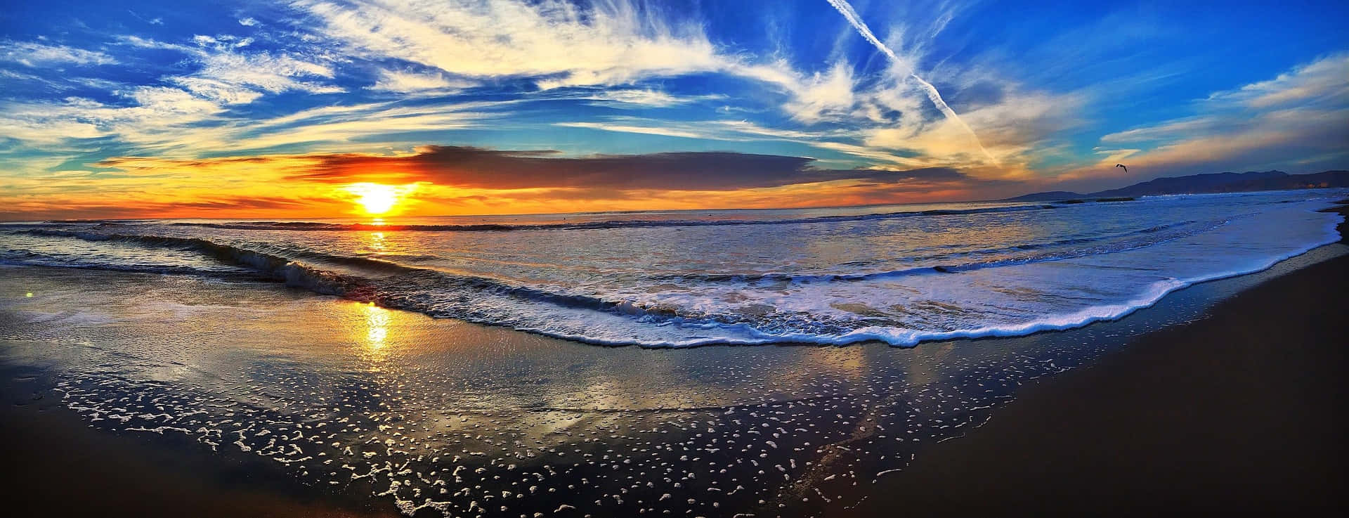 Panoramic 桌布 壁紙 — 廣闊海洋上壯麗的日落，金色波浪拍打著深色沙灘，湛藍天空點綴著縷縷薄雲。