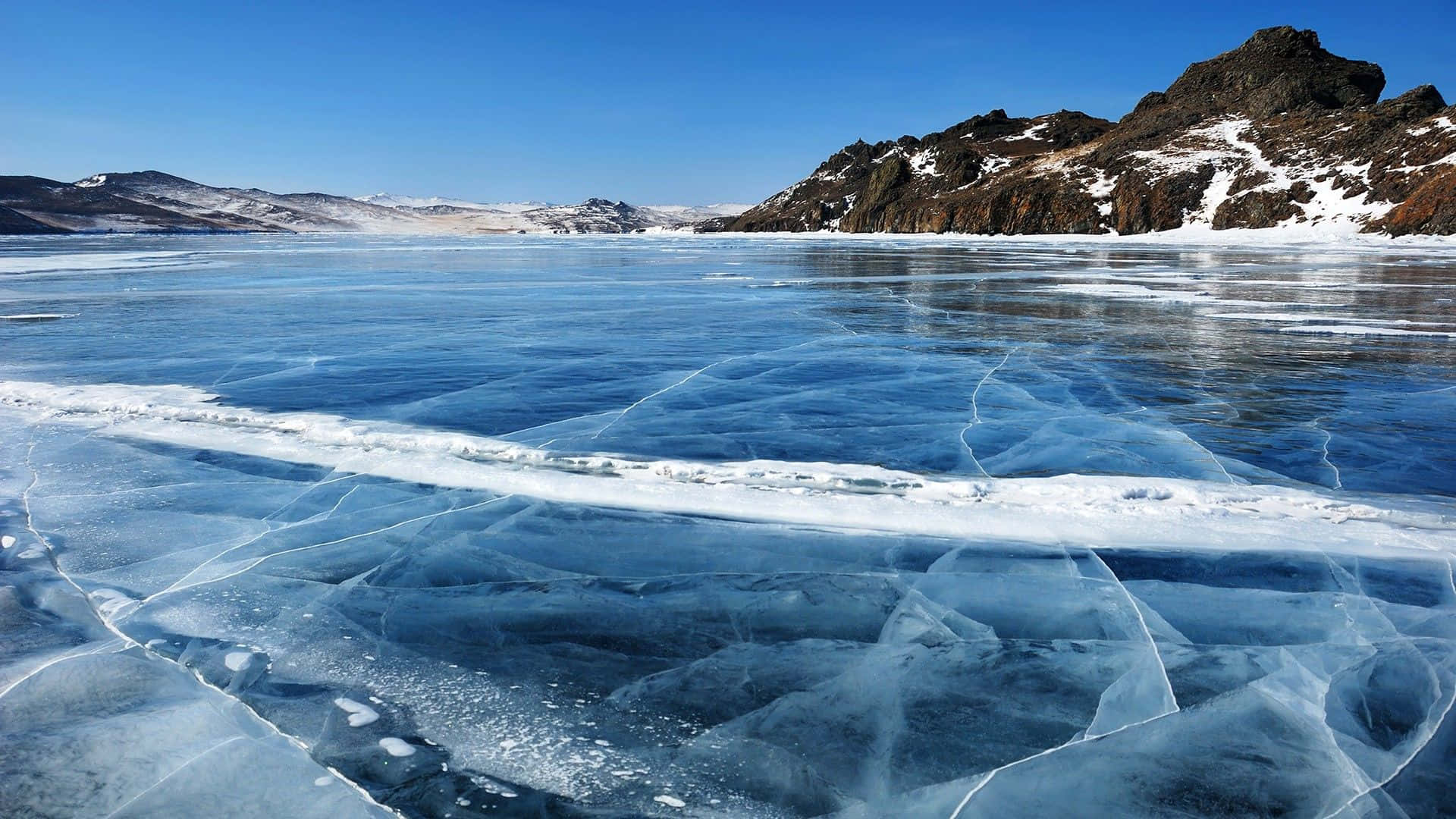 Frozen Lake 桌布 壁紙 — 廣闊的冰凍湖泊，在清澈的藍天下呈現複雜的藍色冰紋，遠處有積雪的山巒。