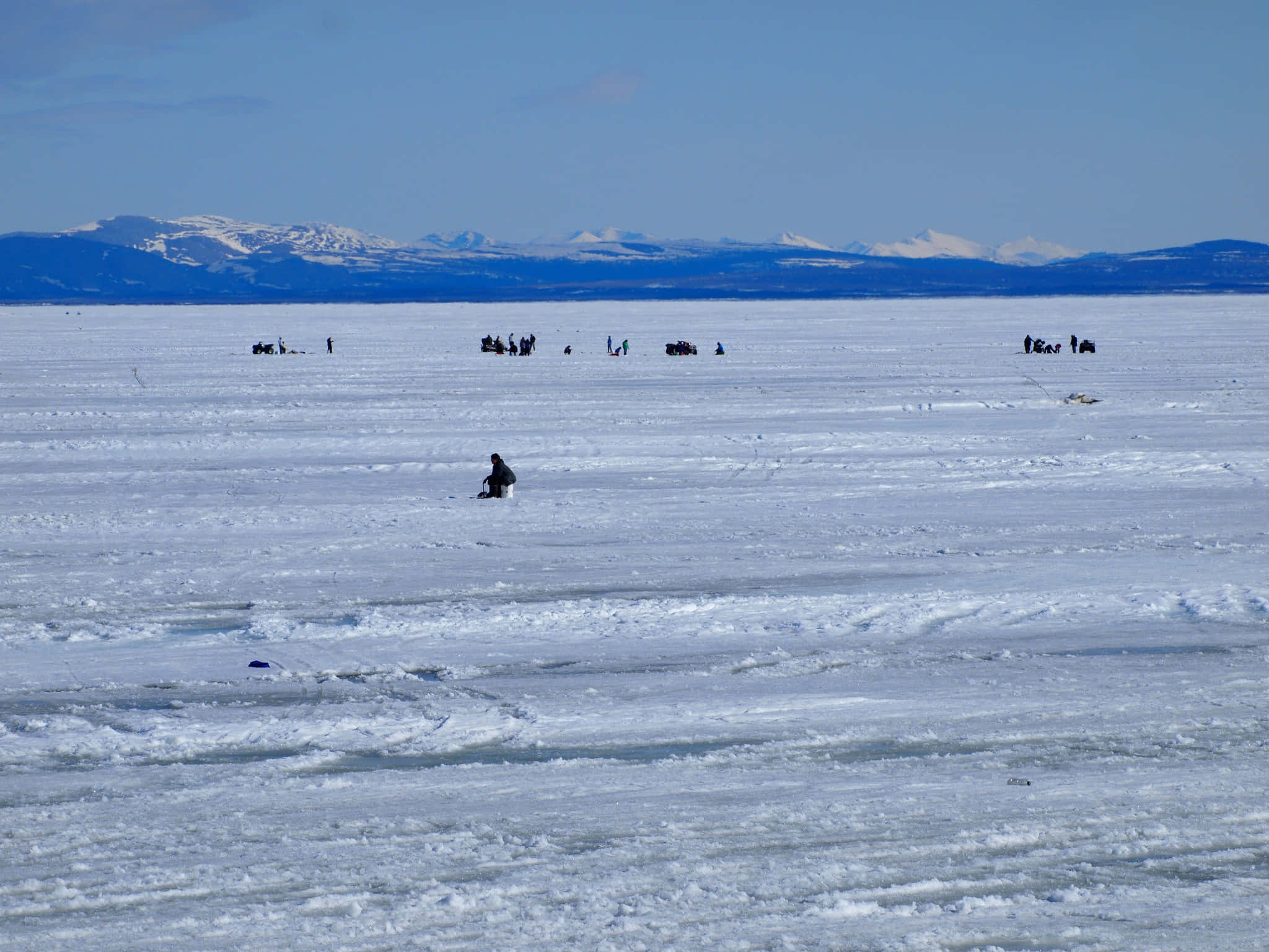 Ice Fishing 桌布 壁紙 — 廣闊的冰凍湖泊，清澈藍天下遠方有積雪山脈，散落著冰釣者。
