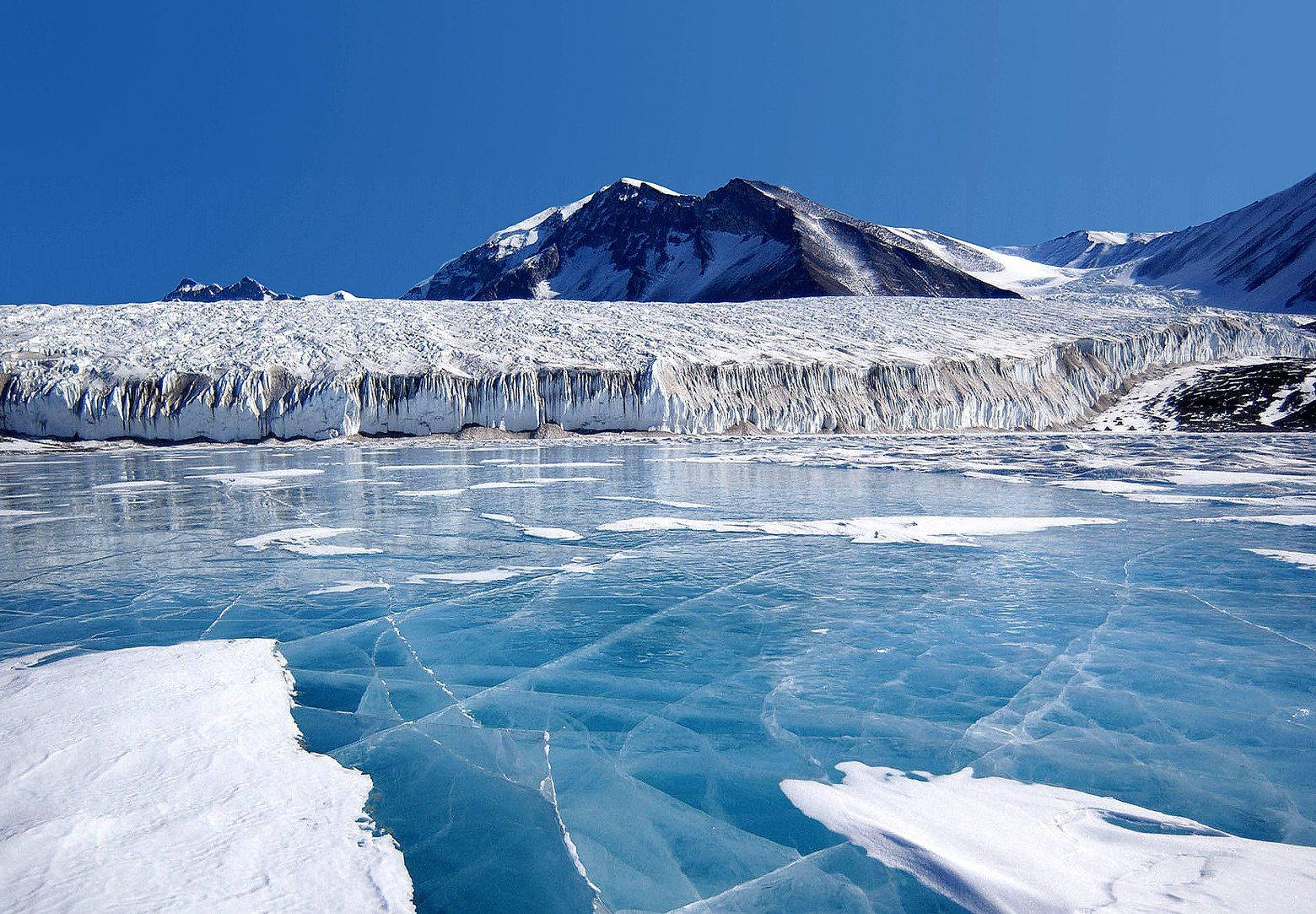 Arctic 桌布 壁紙 — 廣闊的冰川和參差不齊的冰崖聳立在一個結冰的湖面上，湖面有裂開的藍色冰層，天空清澈湛藍。