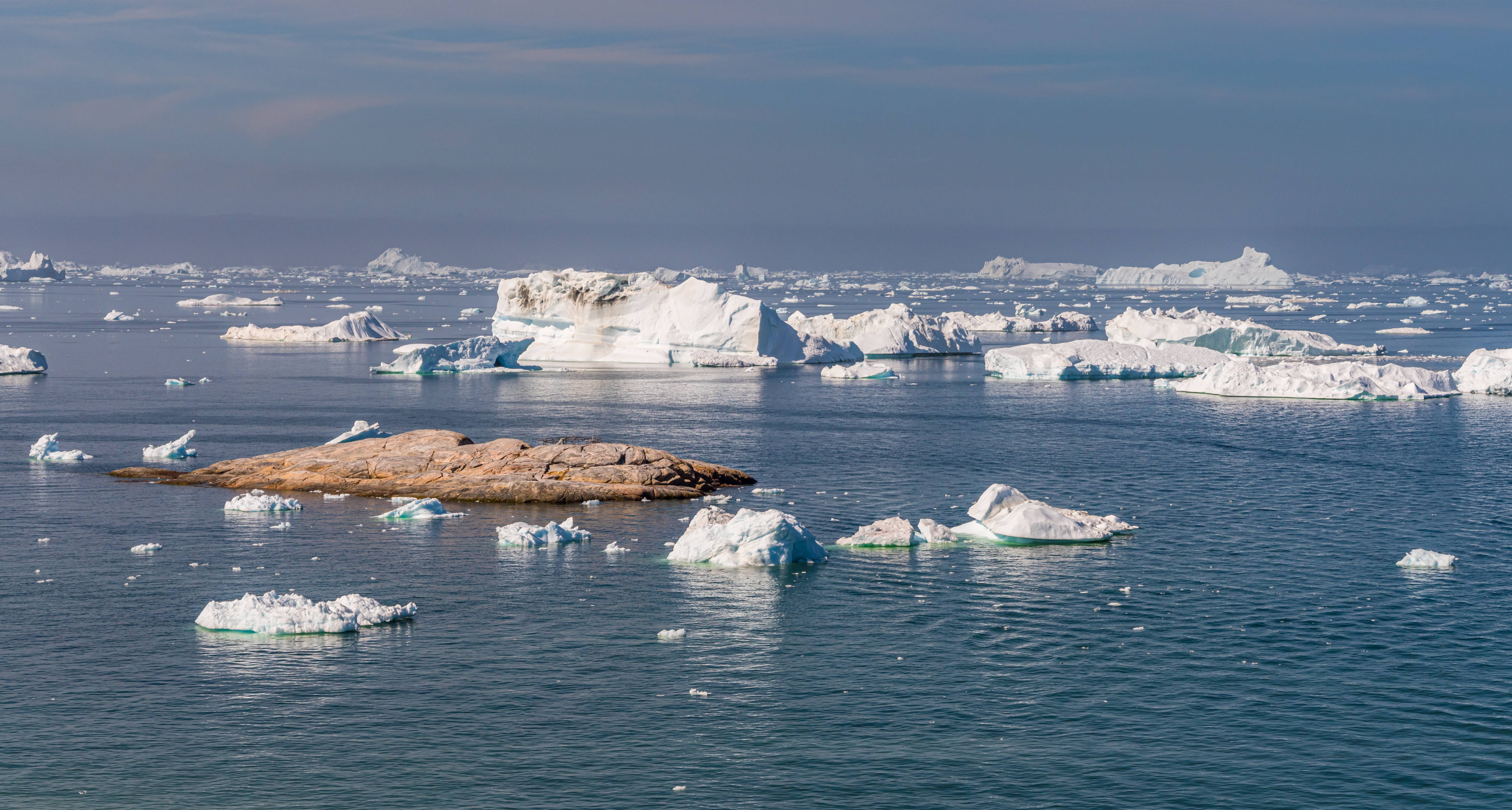Greenland 桌布 壁紙 — 廣闊的北極海景，漂浮的冰山和岩石露頭在淡藍色的天空下。