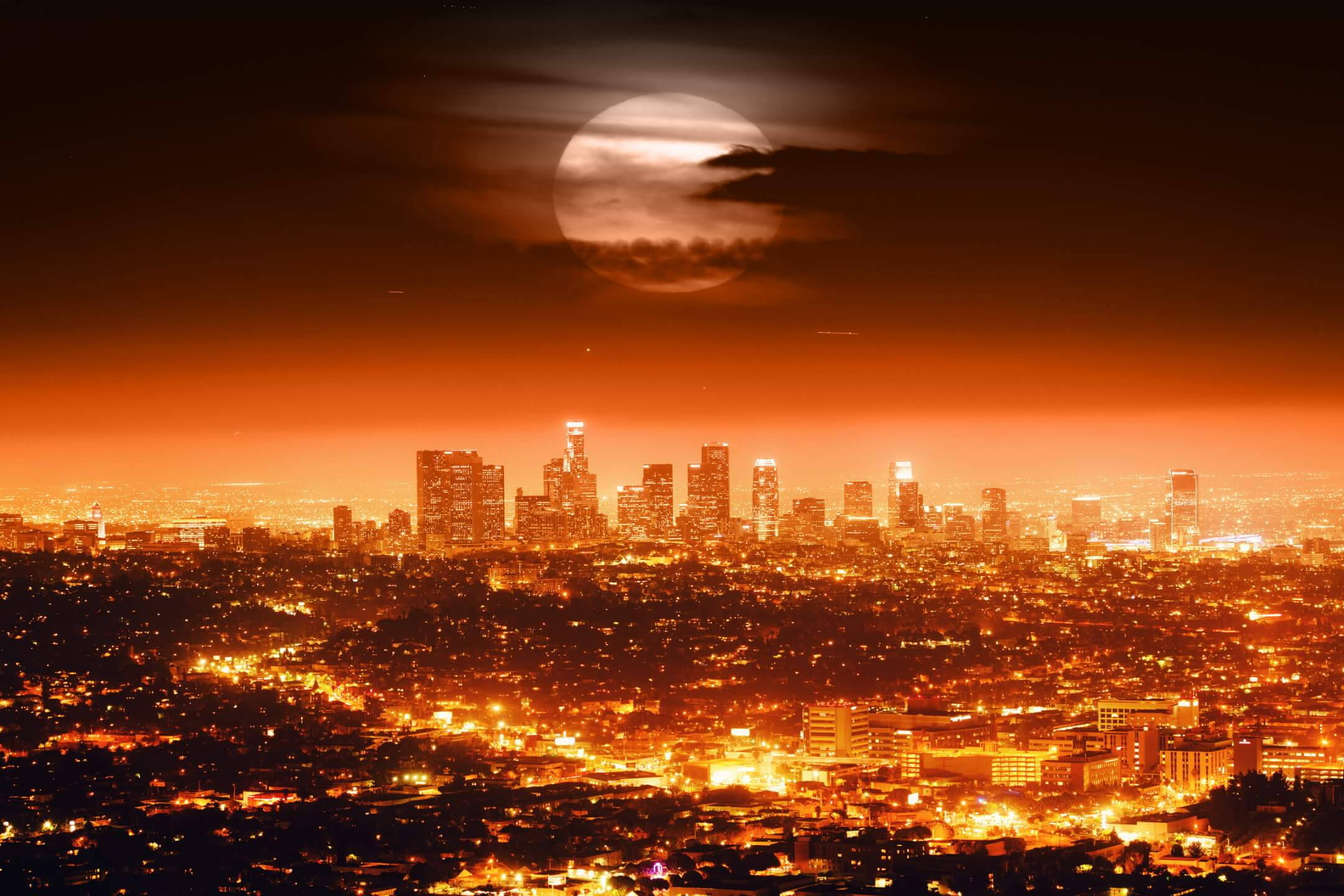 圖片 桌布 壁紙 — 廣闊的城市夜景，在溫暖的橙色光芒中，一輪巨大的月亮部分被雲層遮蓋。