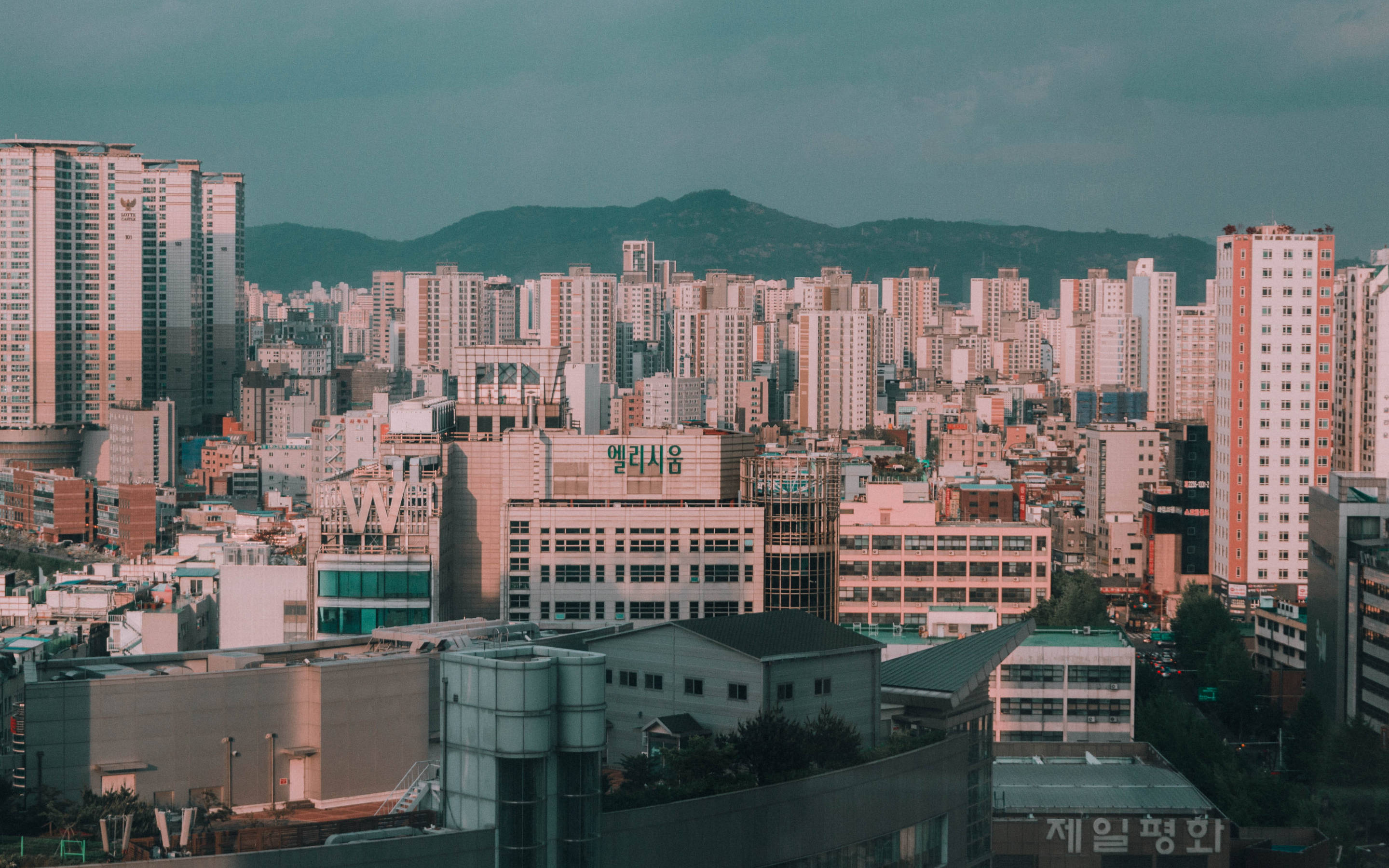 korean 桌布 壁紙 — 廣闊的城市天際線，密集的高層建築，在憂鬱的藍綠色天空下，遠山可見。