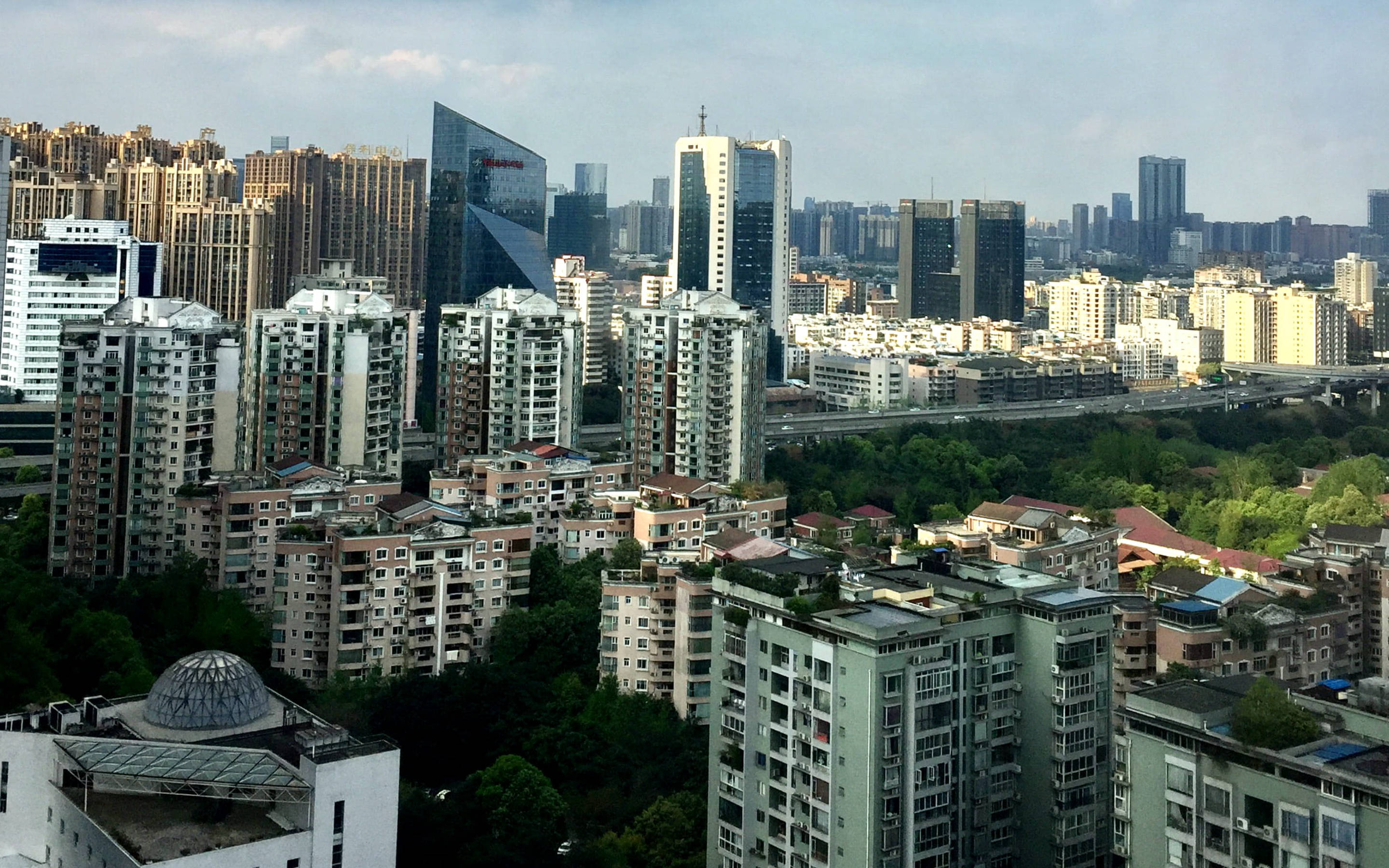 Chengdu 桌布 壁紙 — 廣闊的城市天際線，現代玻璃塔樓聳立在密集的住宅區和茂密的綠樹之上，天空朦朧。