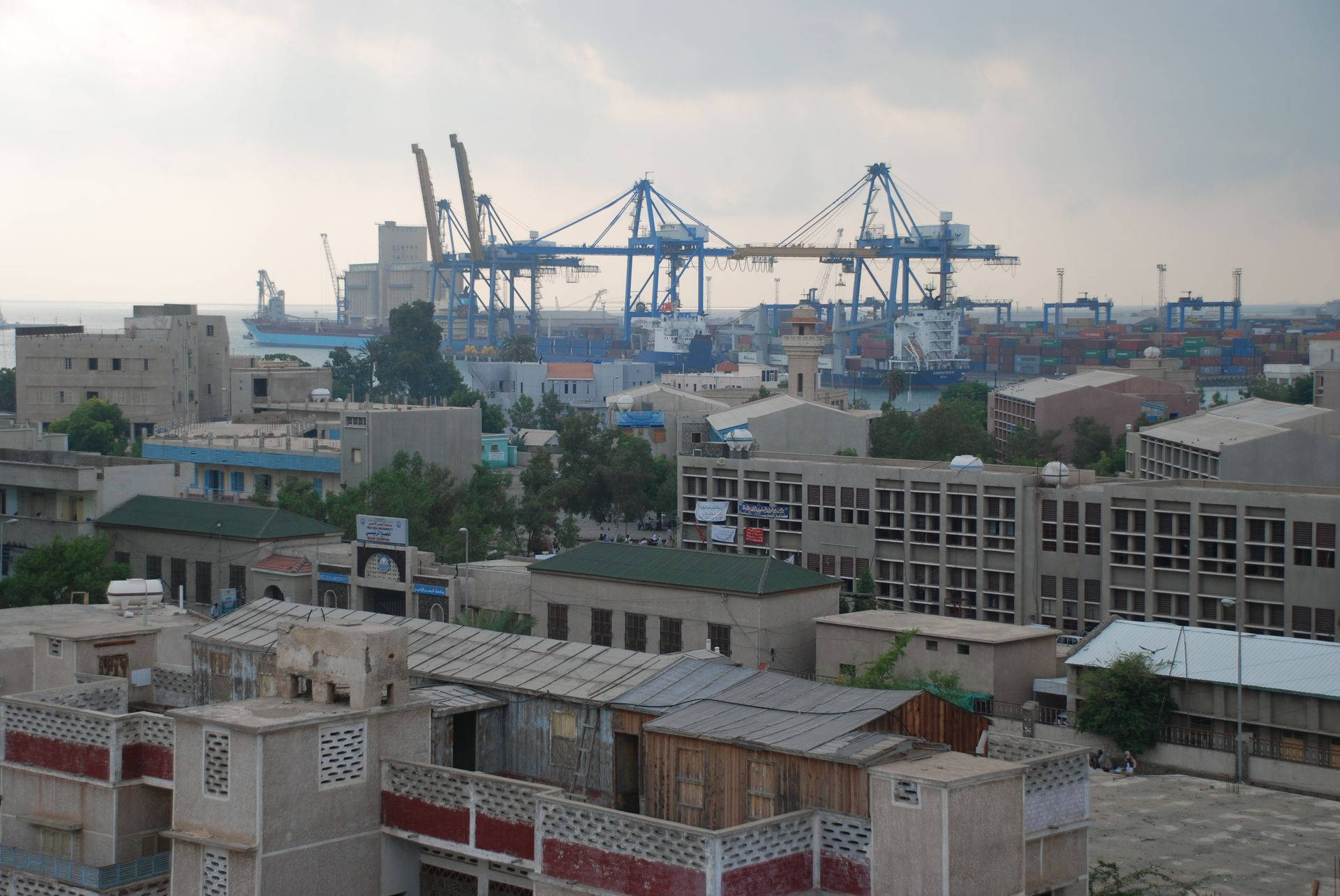 Sudan 桌布 壁紙 — 廣闊的城市景觀，前景是風化的建築物，背景是巨大的藍色港口起重機，在陰天的主宰下。