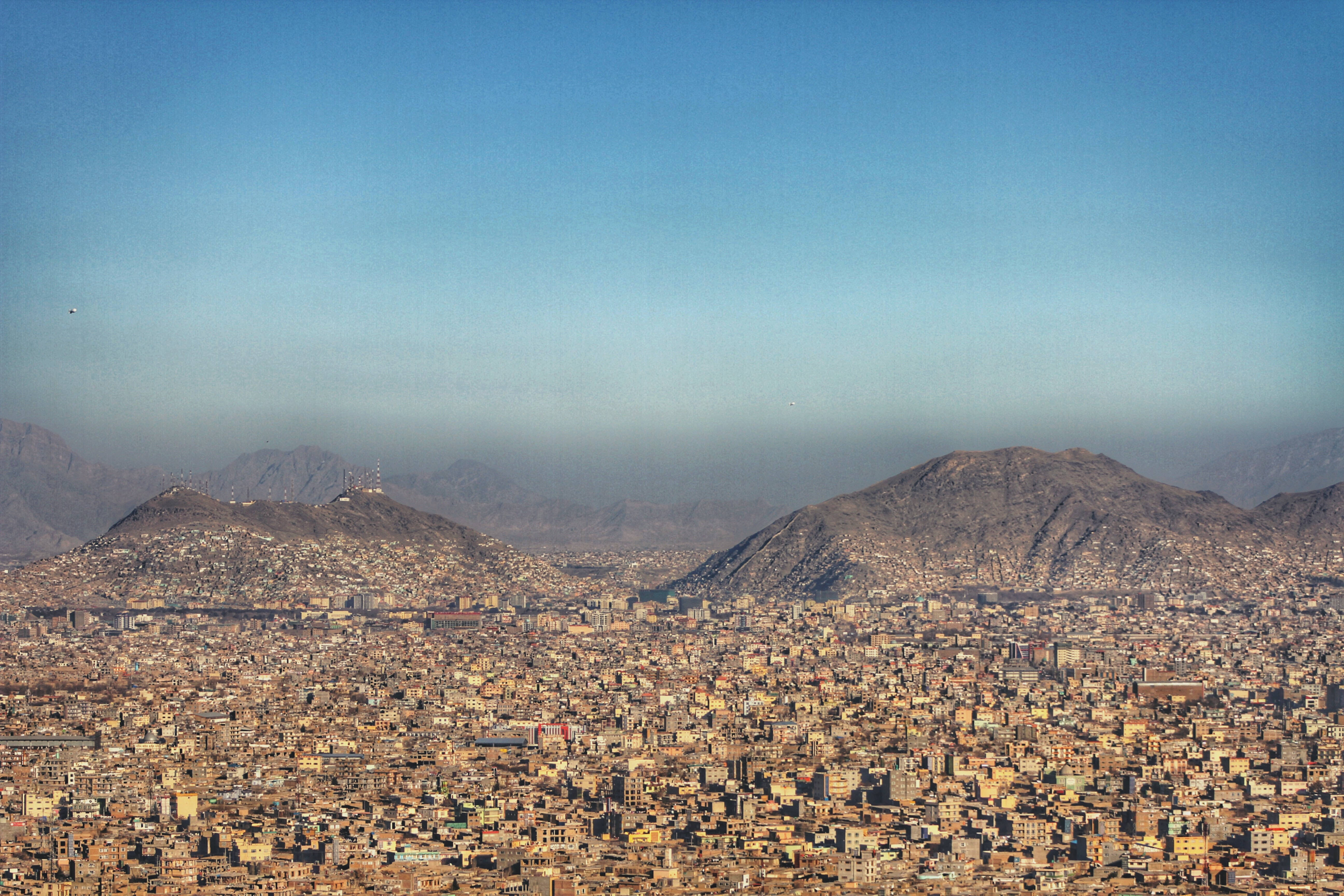 Kabul 桌布 壁紙 — 廣闊的城市景觀，緊密排列的陶土建築群，坐落於崎嶇的棕色山脈之間，上方是清澈的藍天。