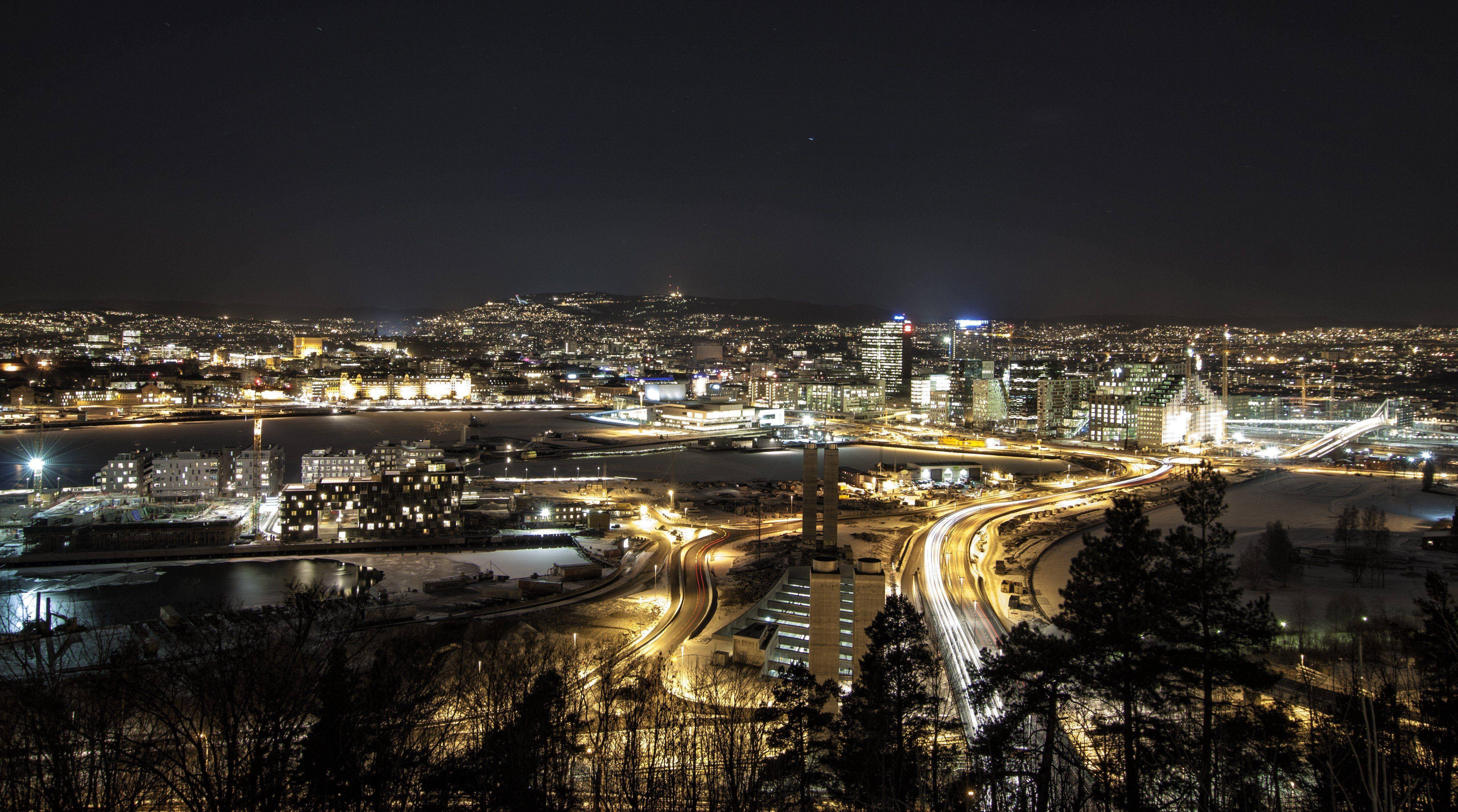 夜空 桌布 壁紙 — 廣闊的夜景城市景觀，建築物發出閃爍燈光，蜿蜒道路上車流形成光跡，並以深色樹木剪影為框。