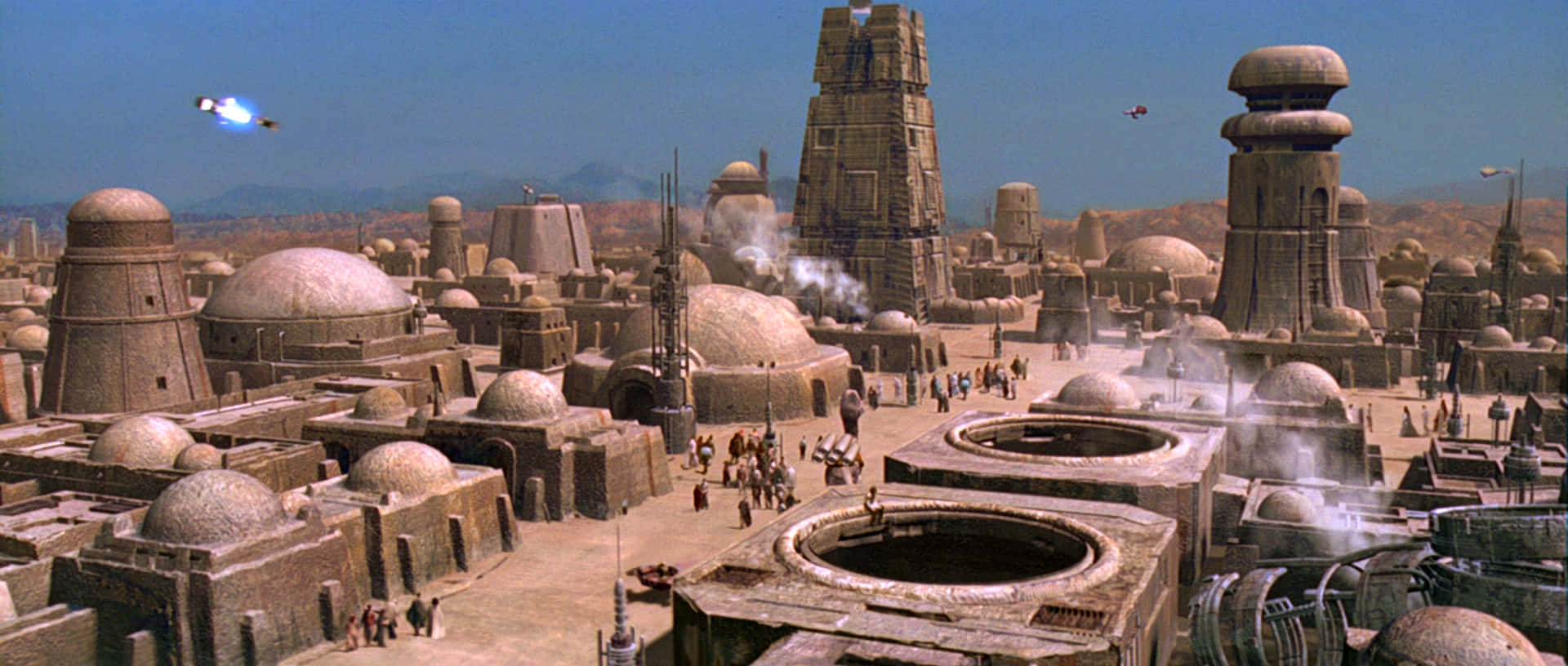 Mos Eisley 桌布 壁紙 — 廣闊的沙漠城市景觀，圓頂建築和高聳的結構在晴朗的藍天下，還有飛行車輛。