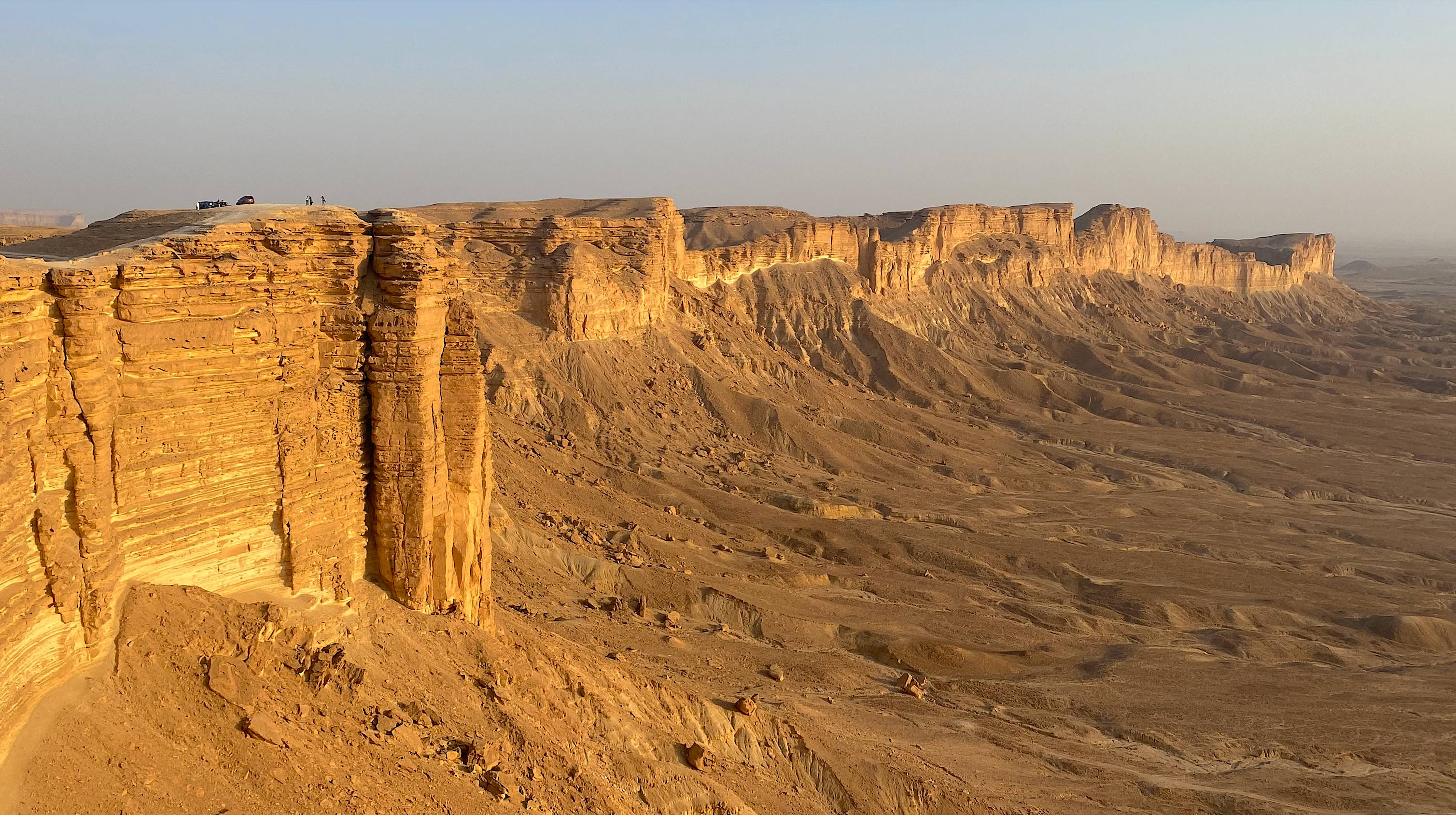 Saudi 桌布 壁紙 — 廣闊的沙漠峽谷，金色懸崖和層狀岩石在晴朗的藍天下。