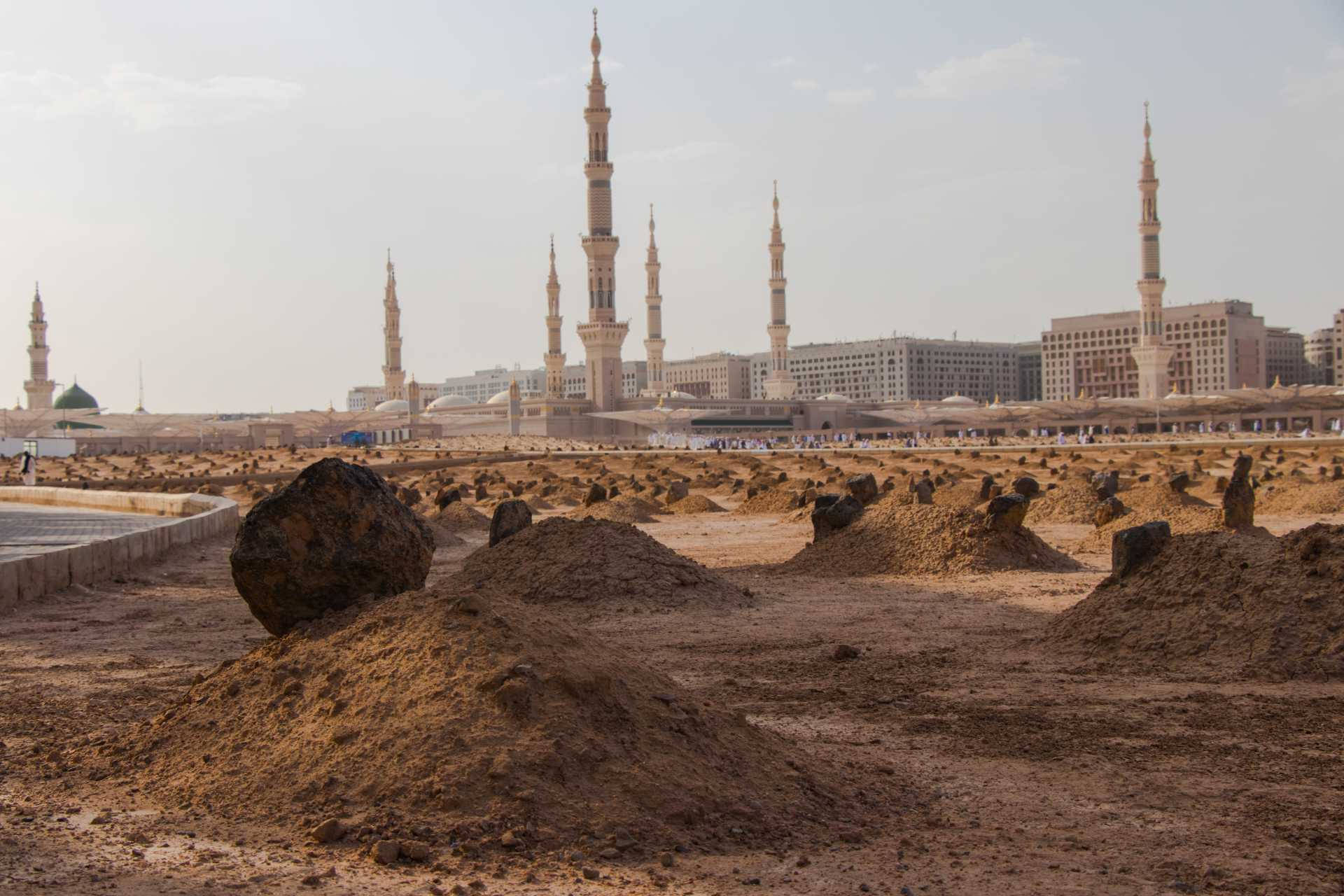 Saudi 桌布 壁紙 — 廣闊的沙漠平原上散佈著土丘和大石，延伸至遠方一座擁有多座纖細宣禮塔的清真寺，天空朦朧。