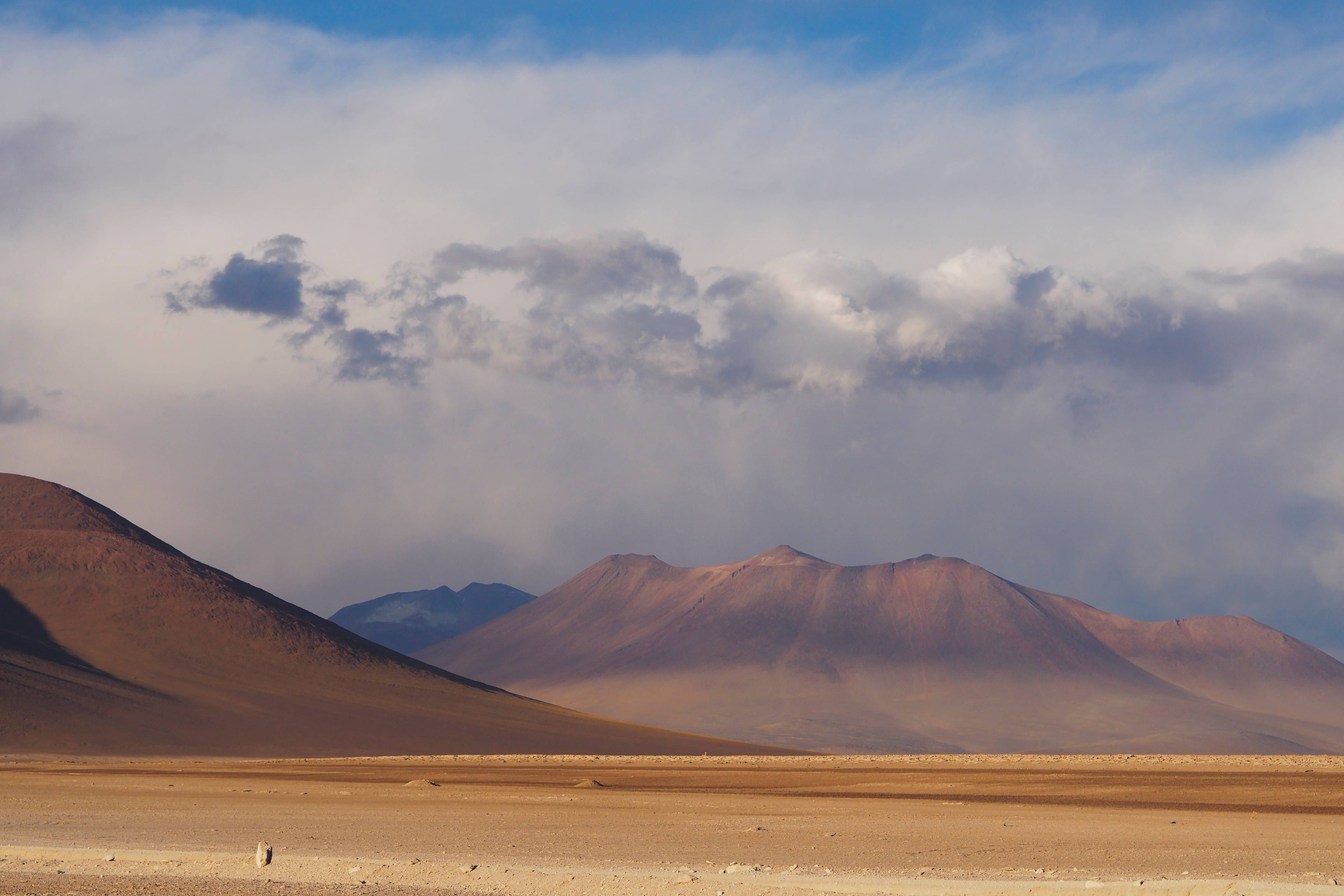 Bolivia 桌布 壁紙 — 廣闊的沙漠平原在戲劇性的天空下延伸，遠處有連綿的微紅色山脈。