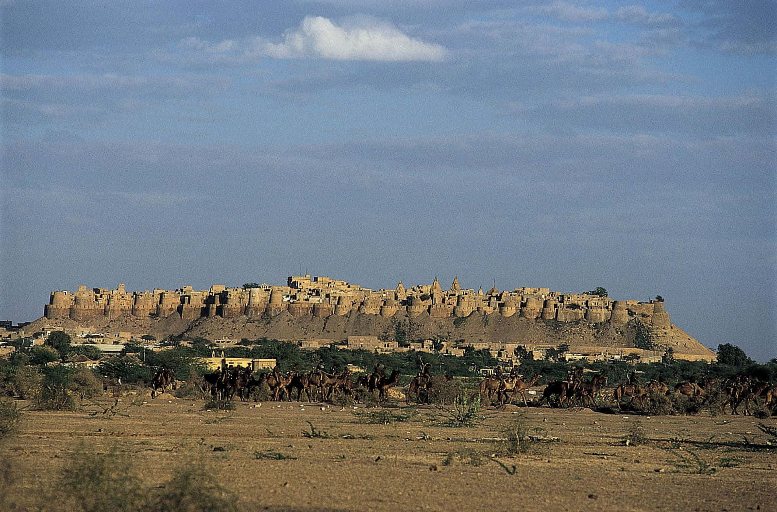 Rajasthan 桌布 壁紙 — 廣闊的沙漠景觀中，一座古老的堡壘矗立在山丘上，一支駱駝商隊在部分多雲的天空下穿越乾燥的前景。