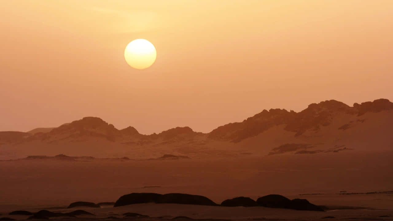 720p Africa 桌布 壁紙 — 廣闊的沙漠景觀沐浴在溫暖的橙色調中，太陽低垂在崎嶇的輪廓山脈之上。