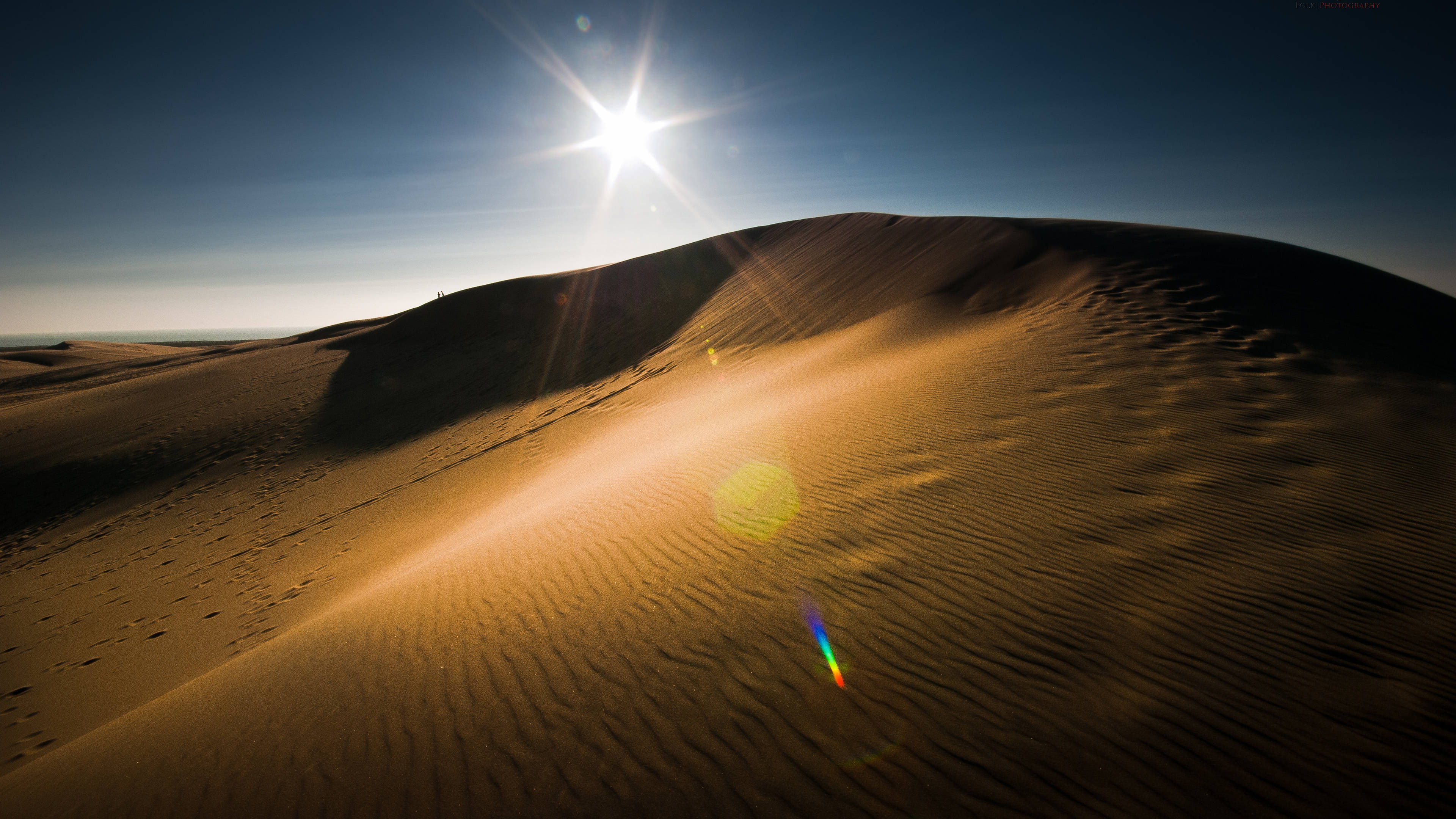 Desert Sun 桌布 壁紙 — 廣闊的沙漠沙丘景觀，沐浴在金色陽光下，鏡頭光暈在深藍色天空下的漣漪沙地上舞動。