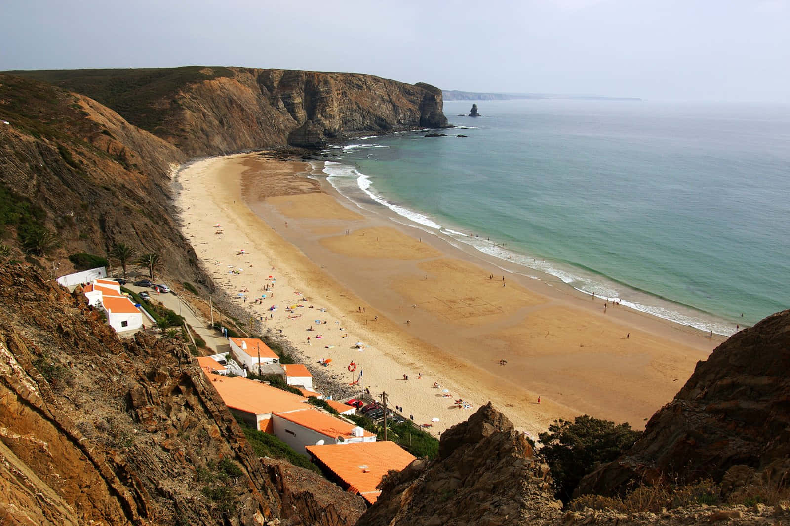Portuguese Beach 桌布 壁紙 — 廣闊的海岸景觀，沙灘坐落在崎嶇的懸崖之間，橘色屋頂的房子緊貼在山坡上。