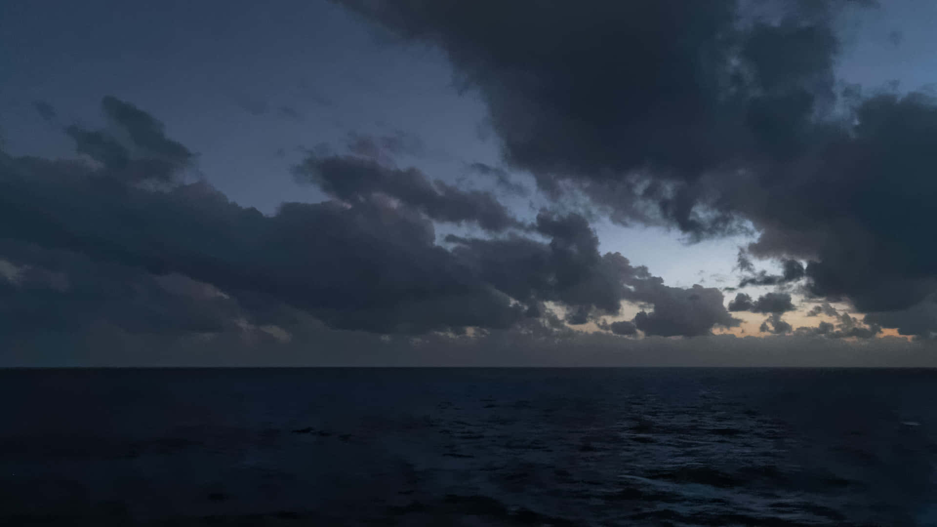 Beautiful Photography 桌布 壁紙 — 廣闊的海洋，天空戲劇性地呈現暮色，烏雲密佈，地平線上有微弱的橙色光芒。
