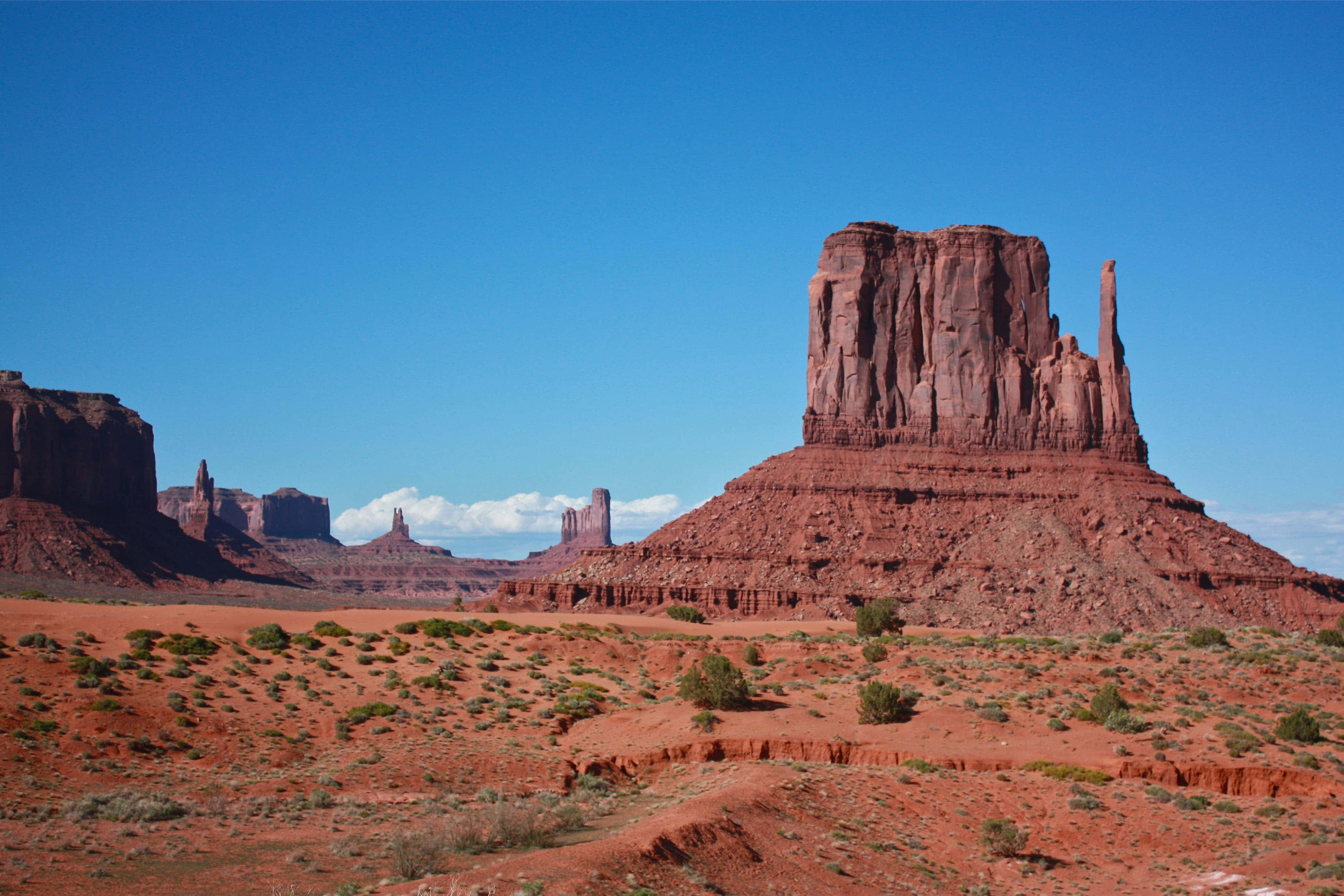 Arizona 桌布 壁紙 — 廣闊的紅色岩層從乾燥的沙漠地形中戲劇性地升起，在清澈的藍天和零星雲朵下。