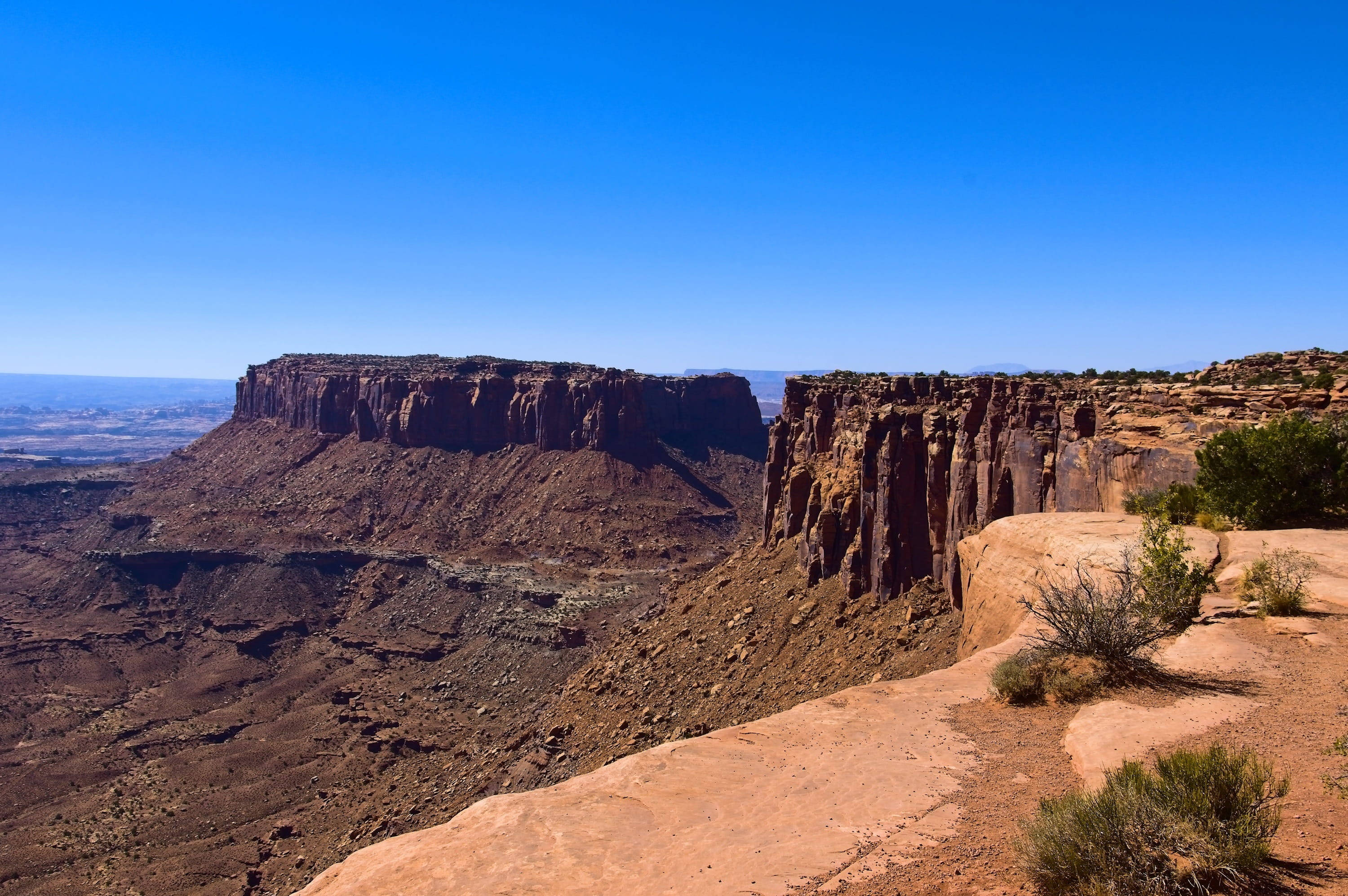 Canyonlands National Park 桌布 壁紙 — 廣闊的紅色岩石峽谷，在清澈的藍天下，有崎嶇的懸崖和稀疏的沙漠植被。