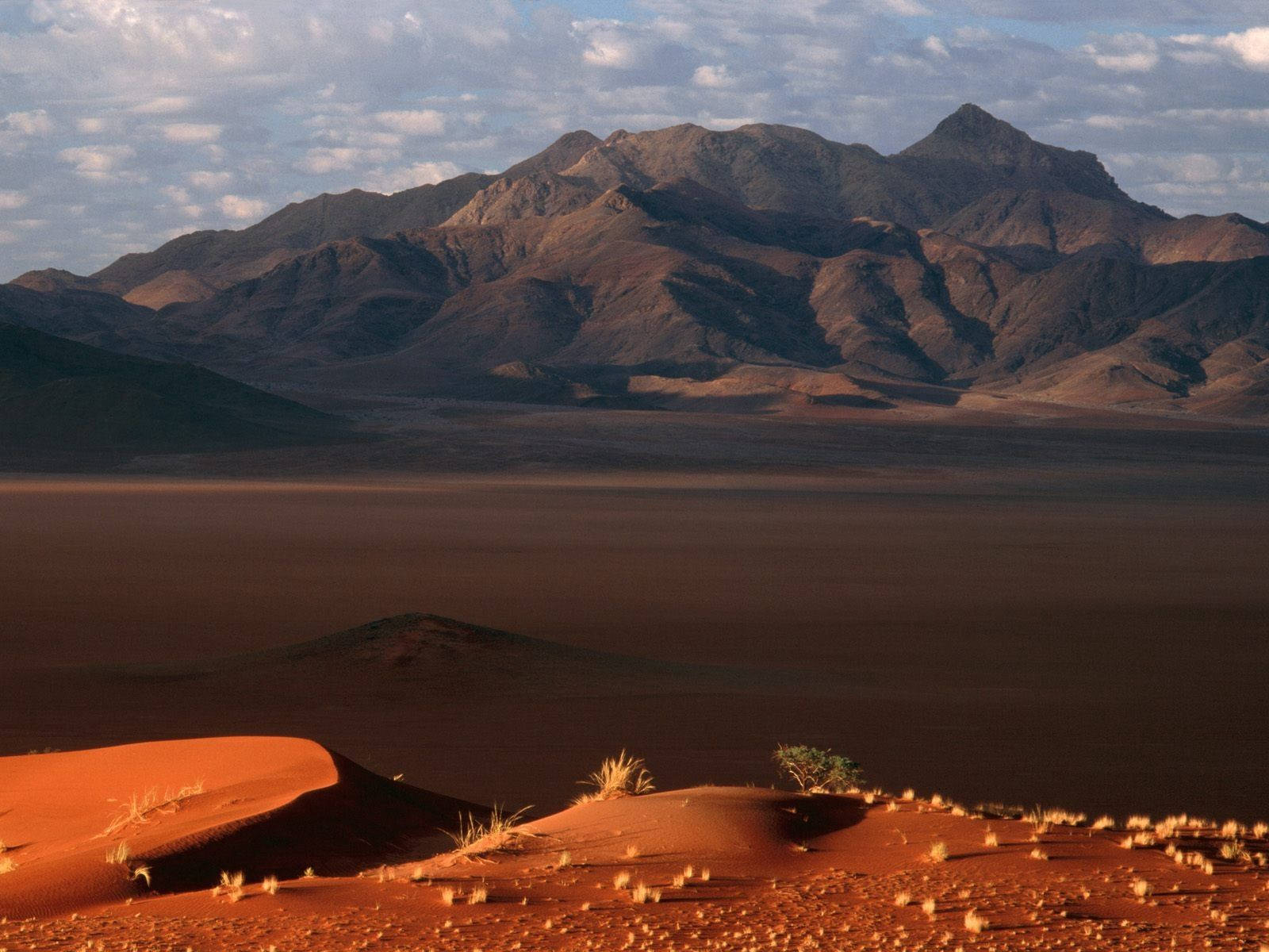Namibia 桌布 壁紙 — 廣闊的紅色沙丘在部分多雲的天空下延伸至崎嶇的山脈，呈現寧靜的沙漠景觀。