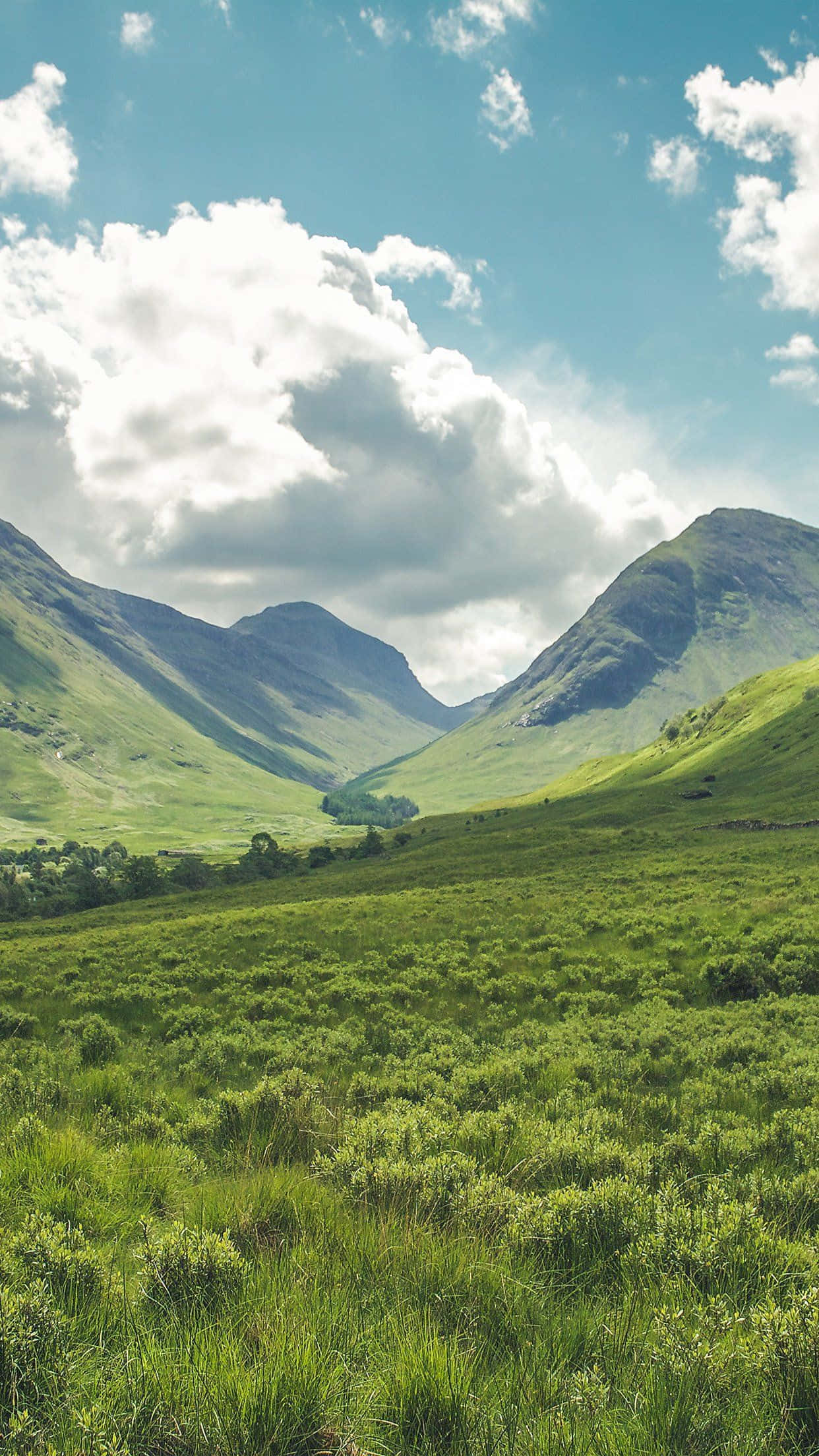Green Nature iPhone 桌布 壁紙 — 廣闊的綠色山谷坐落在崎嶇的山脈之間，上方是明亮的藍天和蓬鬆的白雲。