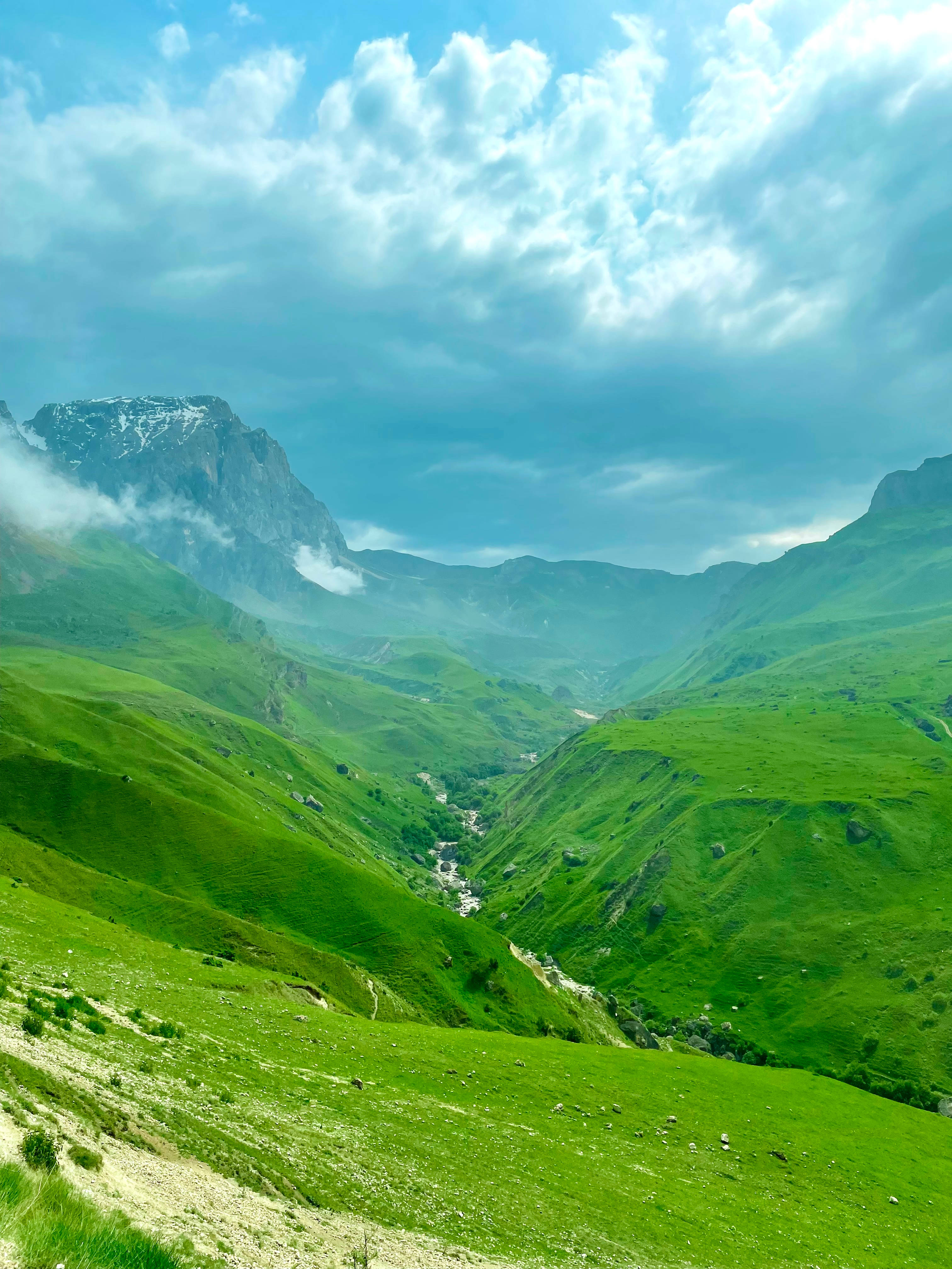 Azerbaijan 桌布 壁紙 — 廣闊的綠色山谷坐落於高聳的山脈之間，天空多雲而戲劇化，遠處有積雪的山峰。