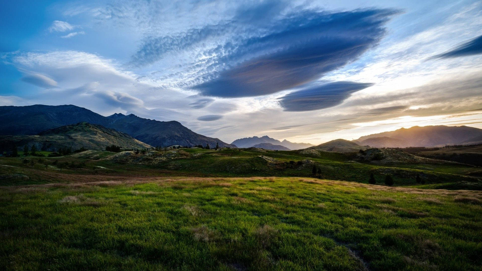 夏天 桌布 壁紙 — 廣闊的綠色草地延伸至遠山，夕陽下天空戲劇性地出現透鏡狀雲。