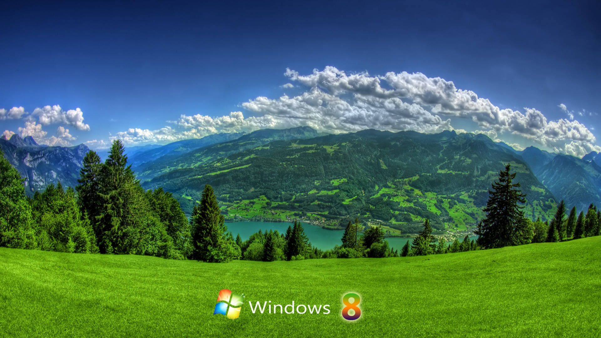 Windows 桌布 壁紙 — 廣闊的綠色草甸向著寧靜的湖泊傾斜，湖泊坐落在高聳的森林覆蓋的山脈之間，上方是明亮的藍天和蓬鬆的白雲。