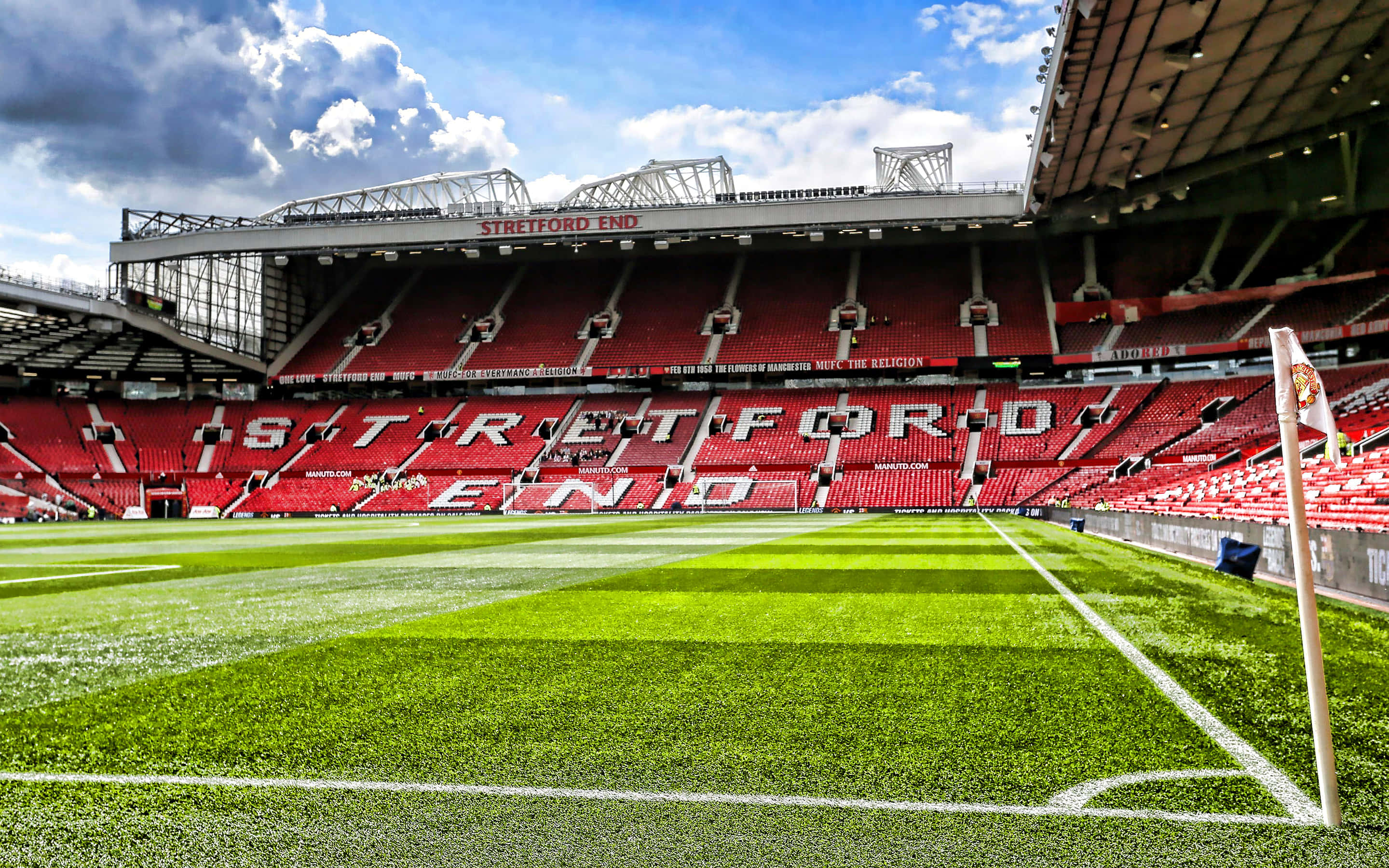 Soccer Stadium 桌布 壁紙 — 廣闊的綠色足球場在戲劇性的多雲天空下，向標誌性的紅色Stretford End看台延伸。