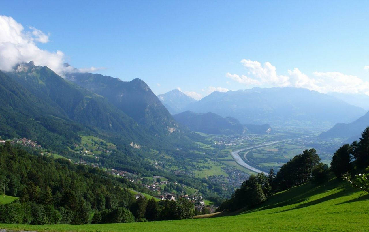 Liechtenstein 桌布 壁紙 — 廣闊的翠綠山谷景觀，坐落於高聳的山脈之間，上方是明亮的藍天，點綴著幾朵白雲。