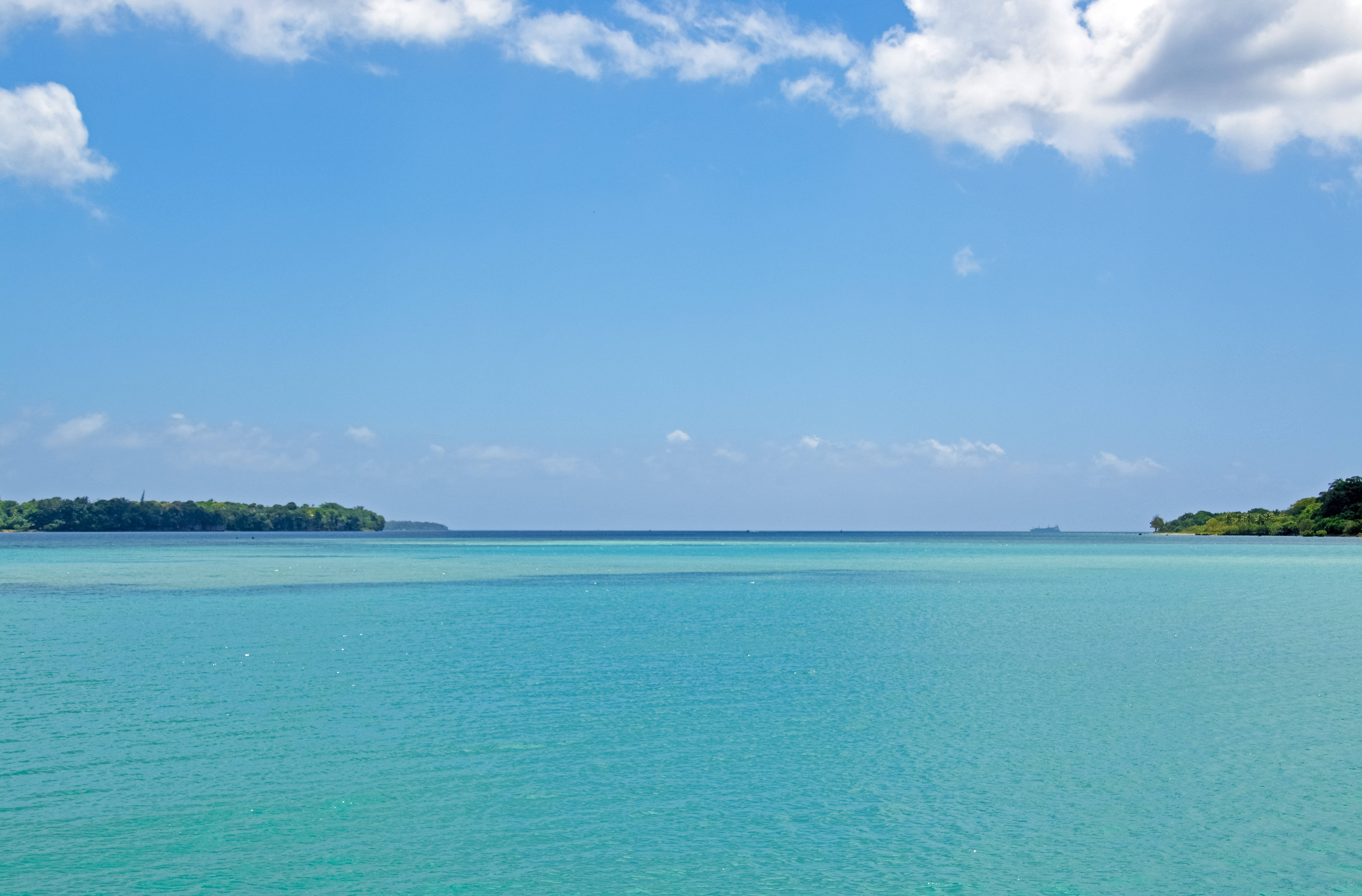 Vanuatu 桌布 壁紙 — 廣闊的藍綠色海洋在明亮的藍天下延伸，散佈著白雲，地平線上有遙遠的綠色島嶼。