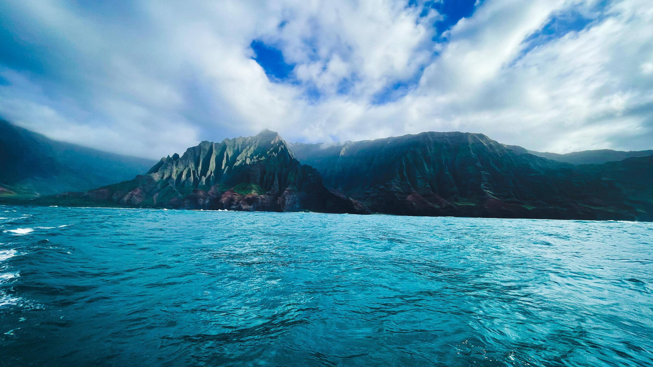 2560x1440 nature 桌布 壁紙 — 廣闊的藍綠色海浪向著戲劇性的鋸齒狀懸崖翻滾，天空湛藍，雲朵散落。
