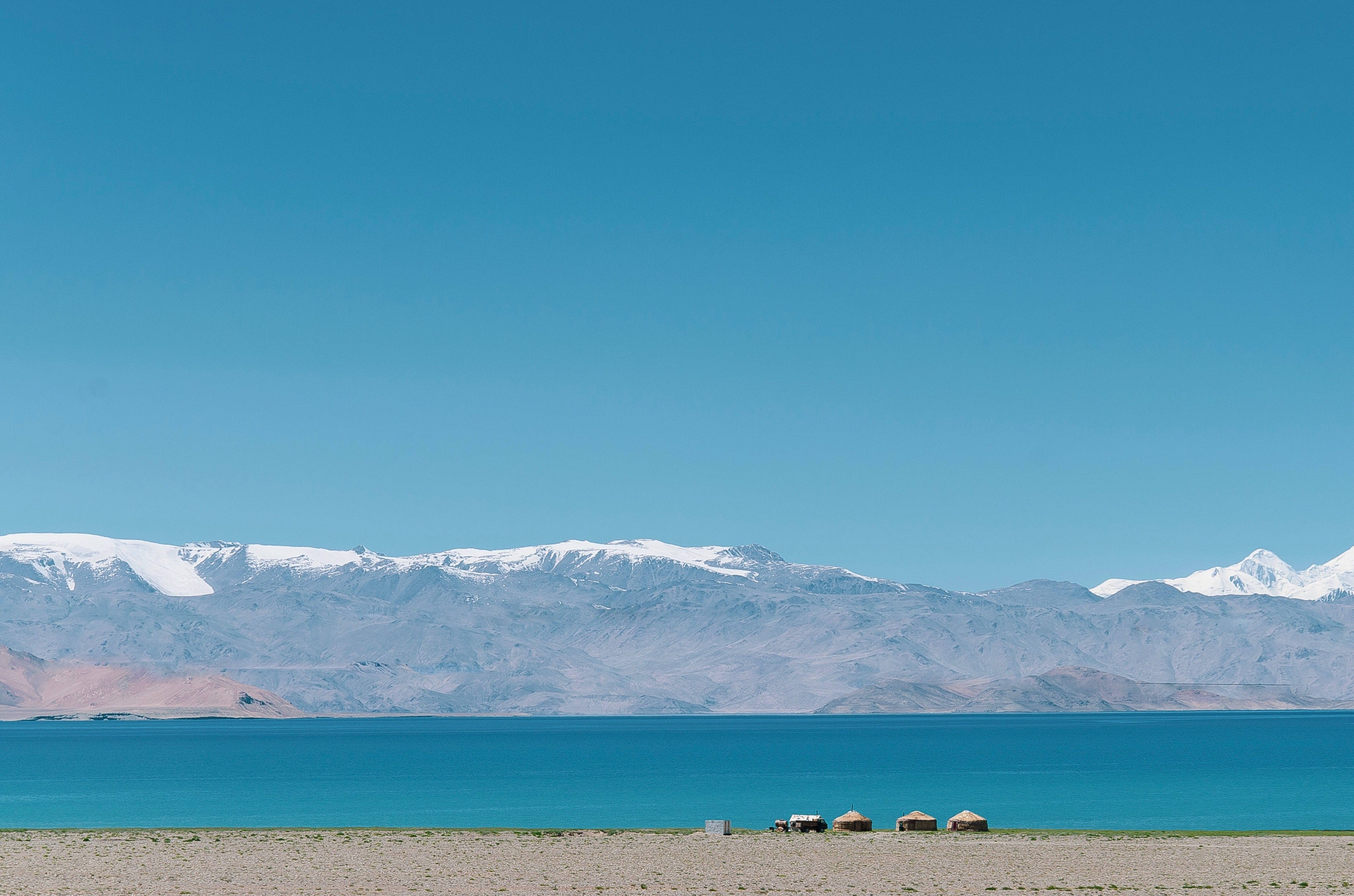 Tajikistan 桌布 壁紙 — 廣闊的藍綠色湖泊，四周環繞著白雪覆蓋的山脈，天空清澈湛藍，岸邊有細小的氈帳。