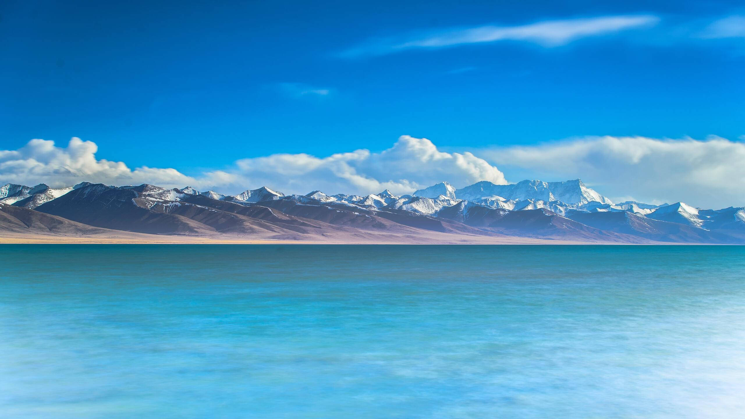 山景 桌布 壁紙 — 廣闊的藍綠色湖泊在白雪覆蓋的山脈前延伸，明亮的藍天下有蓬鬆的白雲。