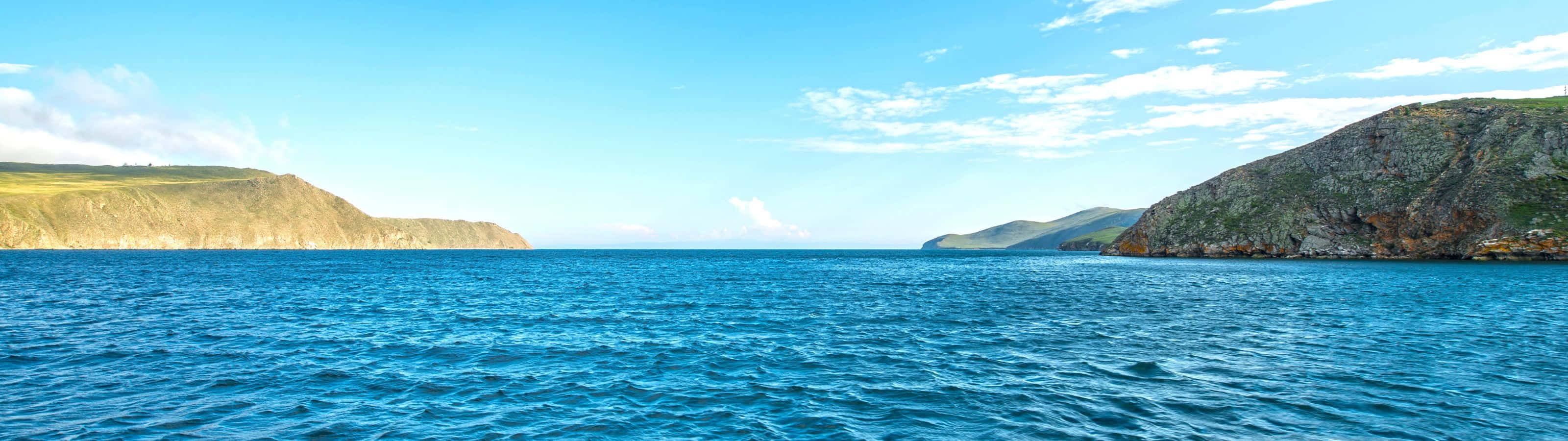 3200x900 桌布 壁紙 — 廣闊的藍色海洋在崎嶇的綠色和棕色懸崖之間延伸，天空明亮清澈，雲朵零星分佈。