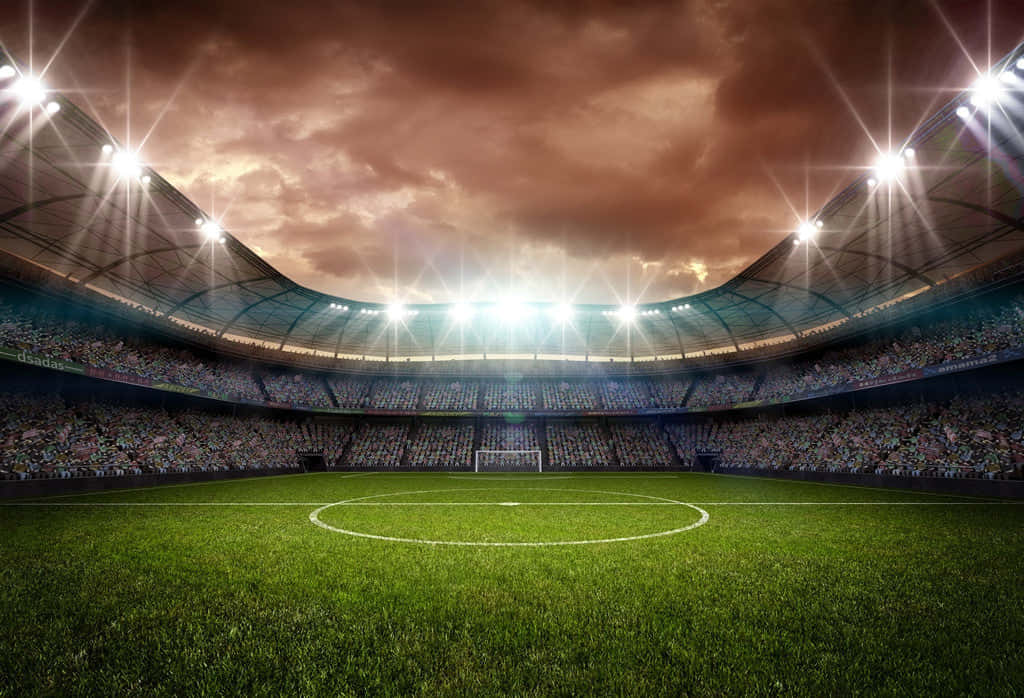 Soccer Stadium 桌布 壁紙 — 廣闊的足球場，在戲劇性的暮色天空下，明亮的泛光燈照亮了翠綠的草地和擠滿觀眾的看台。