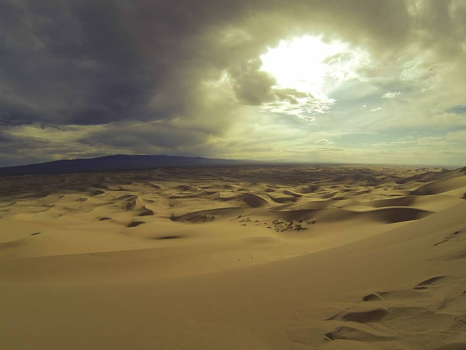 Desert 桌布 壁紙 — 廣闊的金色沙丘在戲劇性的天空下延伸，陽光穿過遠山上的烏雲照射下來。