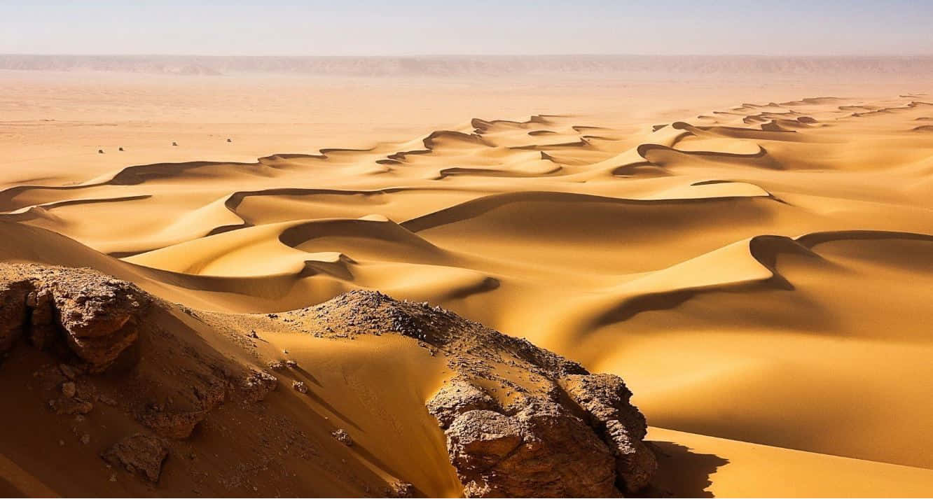 Sand Dunes 桌布 壁紙 — 廣闊的金色沙丘在朦朧的天空下無盡延伸，前景有崎嶇的岩石。