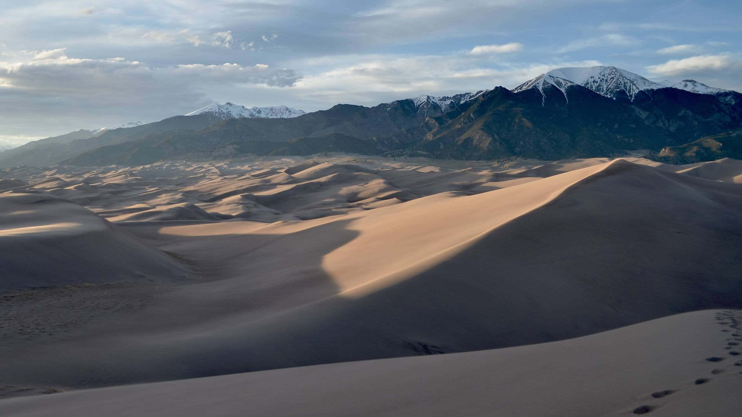 Sand Dunes 桌布 壁紙 — 廣闊的金色沙丘延伸至積雪覆蓋的山脈，天空部分多雲，前景有腳印。