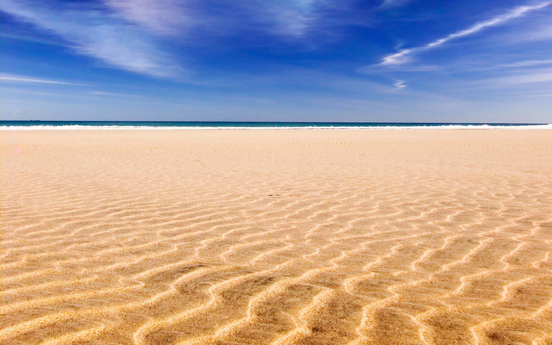 Beach Sand 桌布 壁紙 — 廣闊的金色沙灘帶有波紋紋理，在明亮的藍天和薄雲下延伸至碧綠的海洋。