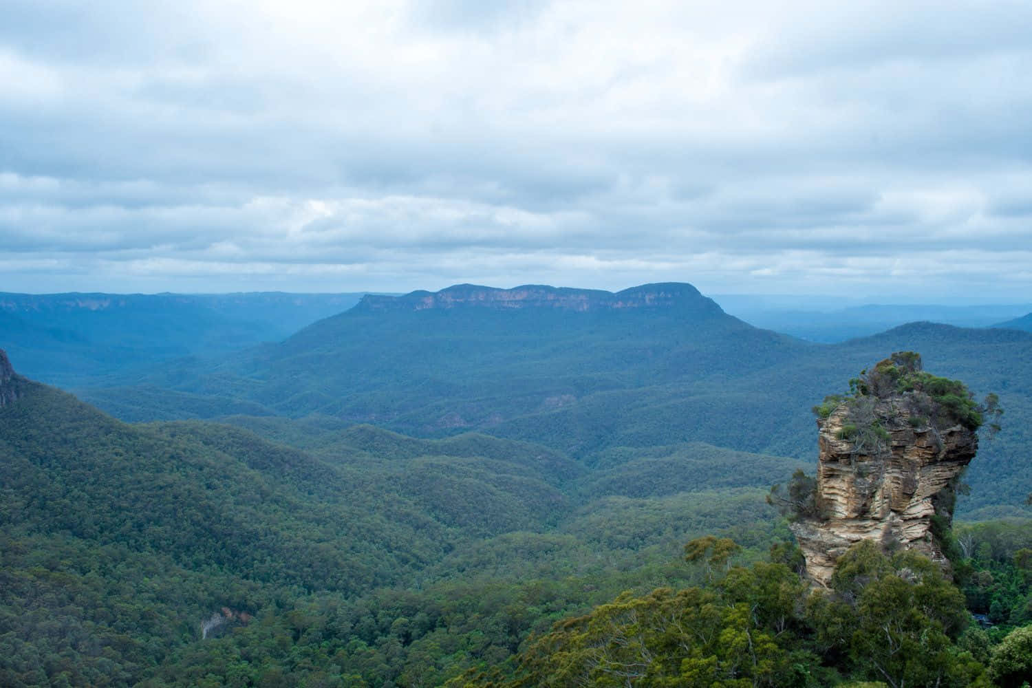 Blue Mountains National Park 桌布 壁紙 — 廣闊的霧谷，層疊的藍綠色山脈，以及一個孤獨的岩石露頭，在陰天天空下覆蓋著綠色植物。