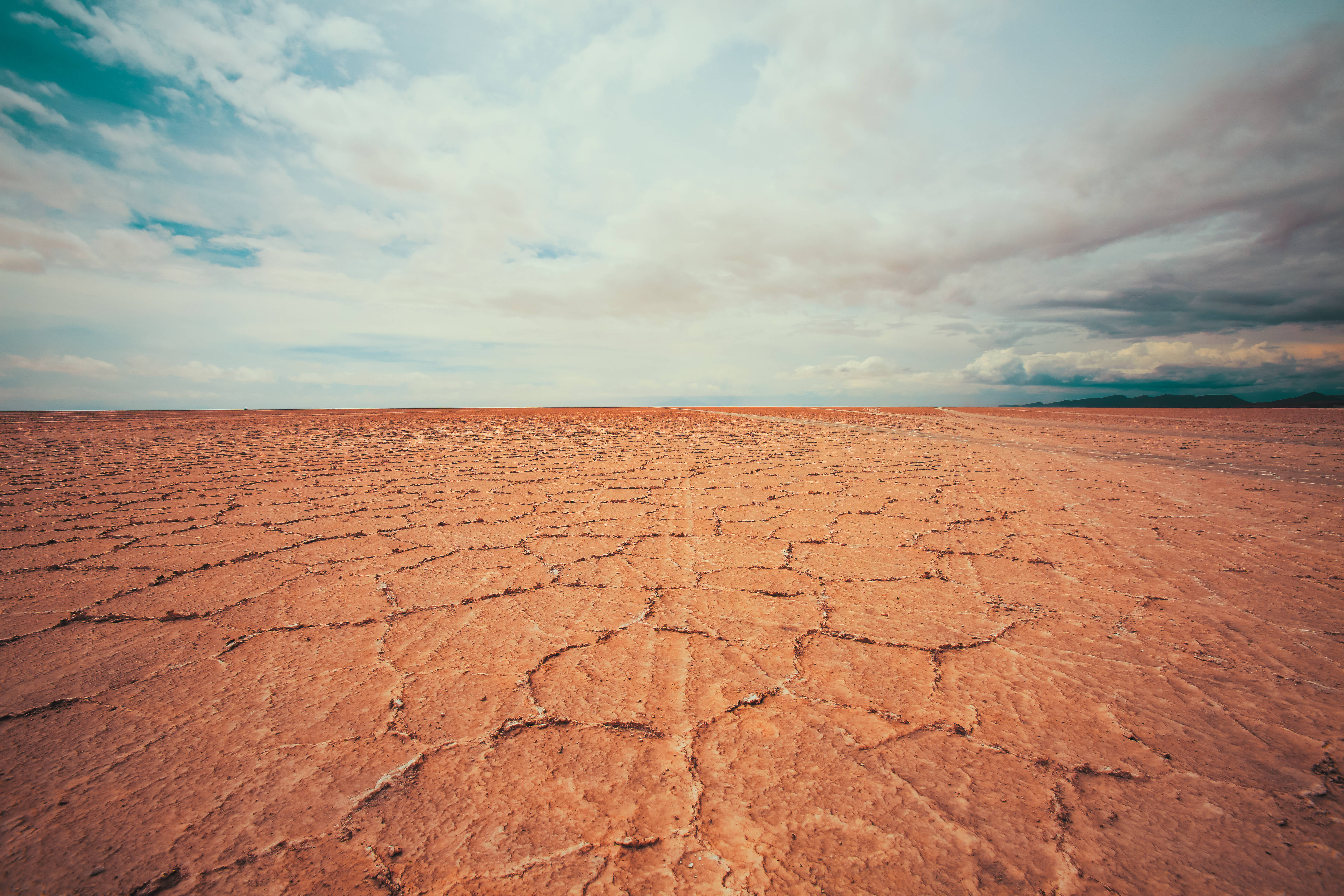 Bolivia 桌布 壁紙 — 廣闊的龜裂大地在戲劇性的天空下延伸，天空中有散落的雲朵和地平線上的遠山。