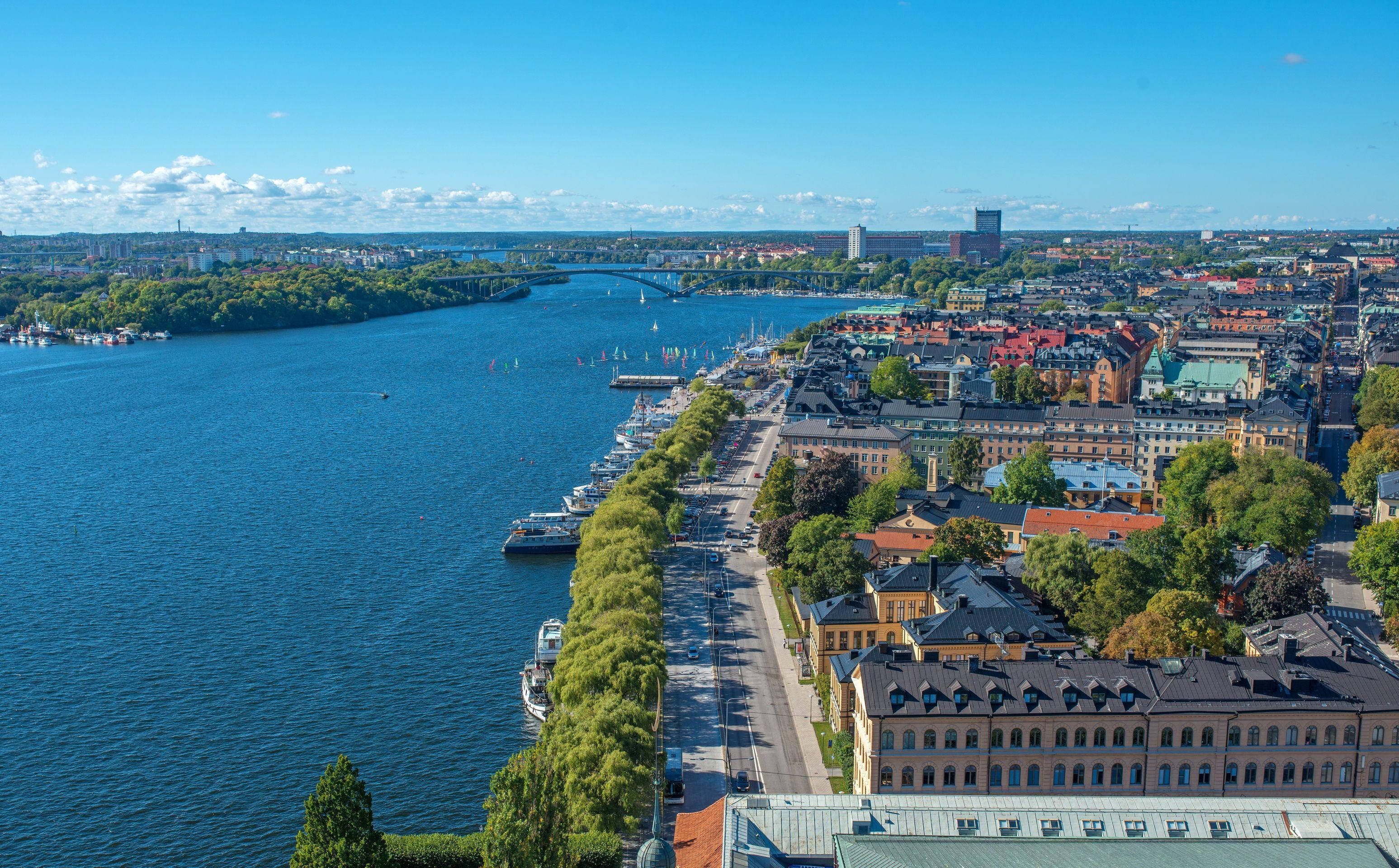 Sweden 桌布 壁紙 — 廣闊藍色河流沿岸充滿活力的城市景觀鳥瞰圖，兩旁種滿茂密的綠色樹木和歷史建築，在晴朗的藍天下。
