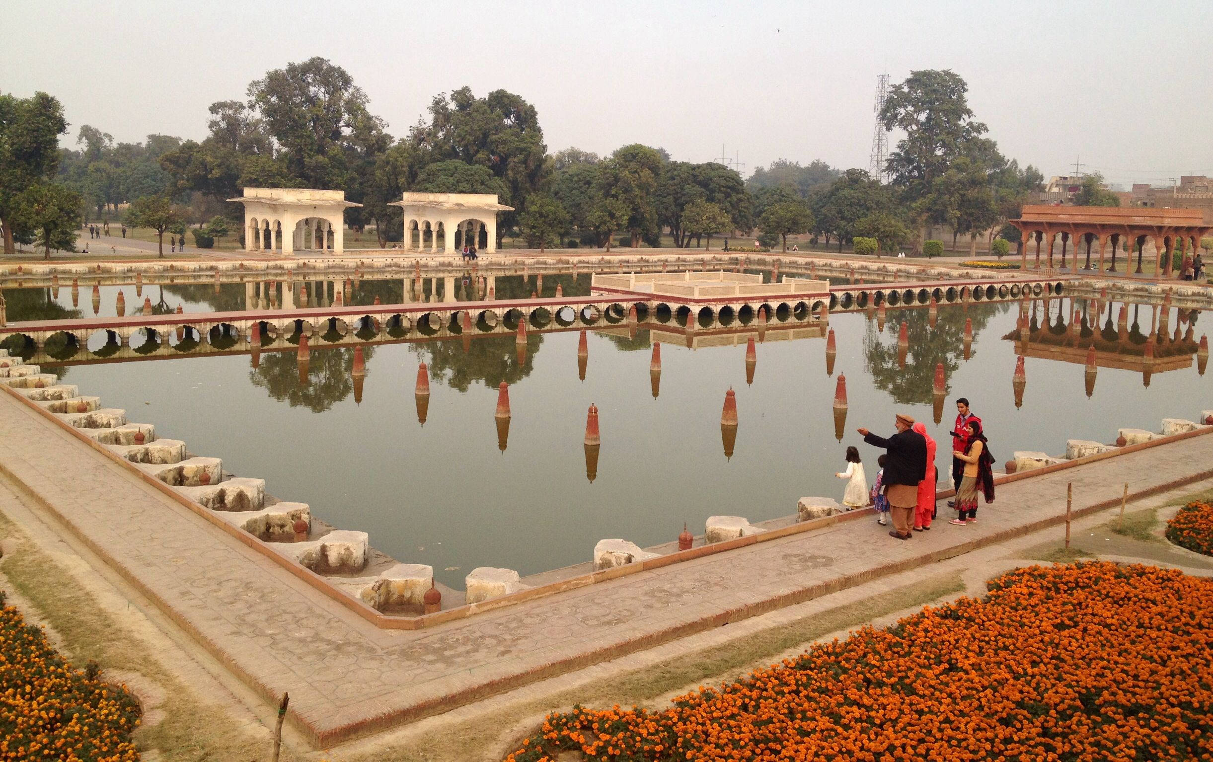 拉合爾 桌布 壁紙 — 廣闊視野的歷史蒙兀兒花園，有一個大型倒影池，周圍環繞著石徑和鮮豔的橙色花壇，天空多雲。