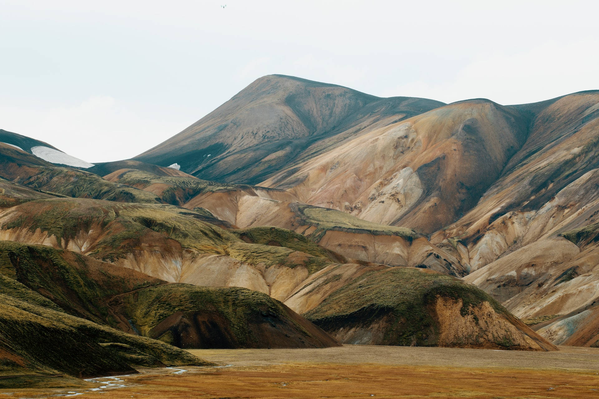 Mountain Landscape 桌布 壁紙 — 廣闊起伏的丘陵，帶有層疊的大地色調和雪斑，天空多雲。